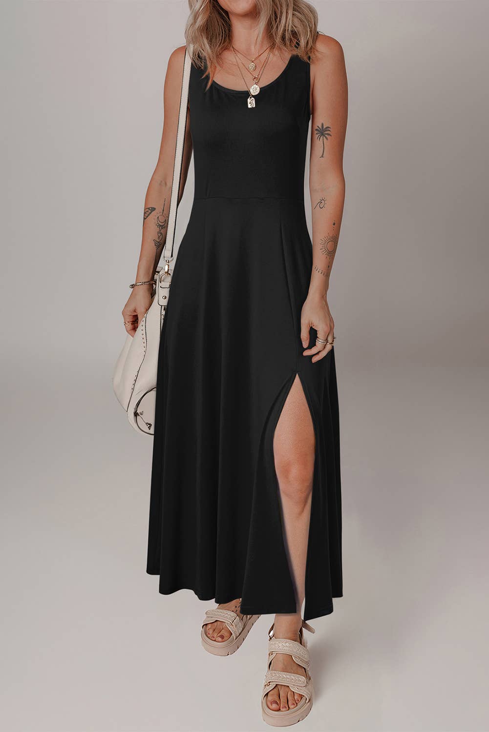 A woman wearing a black solid side split midi dress with no visible sleeves, paired with a white bag and beige footwear. She has accessorized with a necklace.
