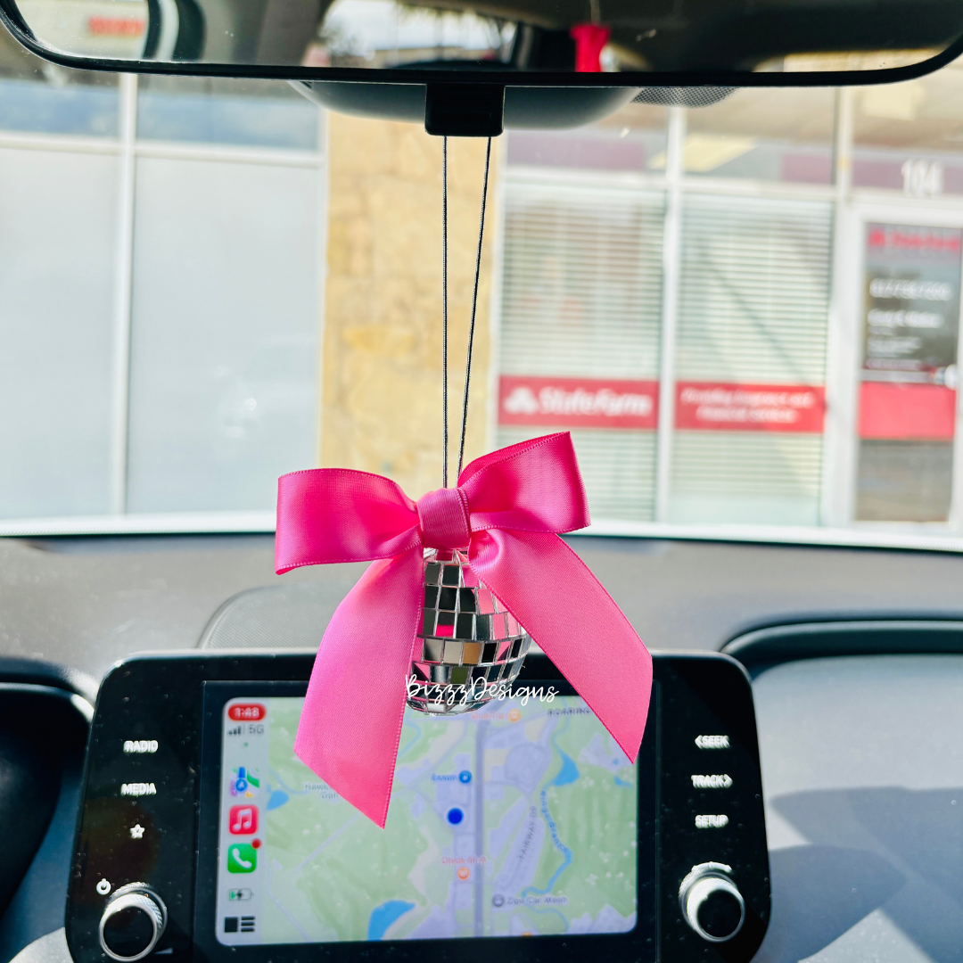 A pink bow disco car charm hanging from a vehicle's rearview mirror featuring a disco ball and pink ribbon.