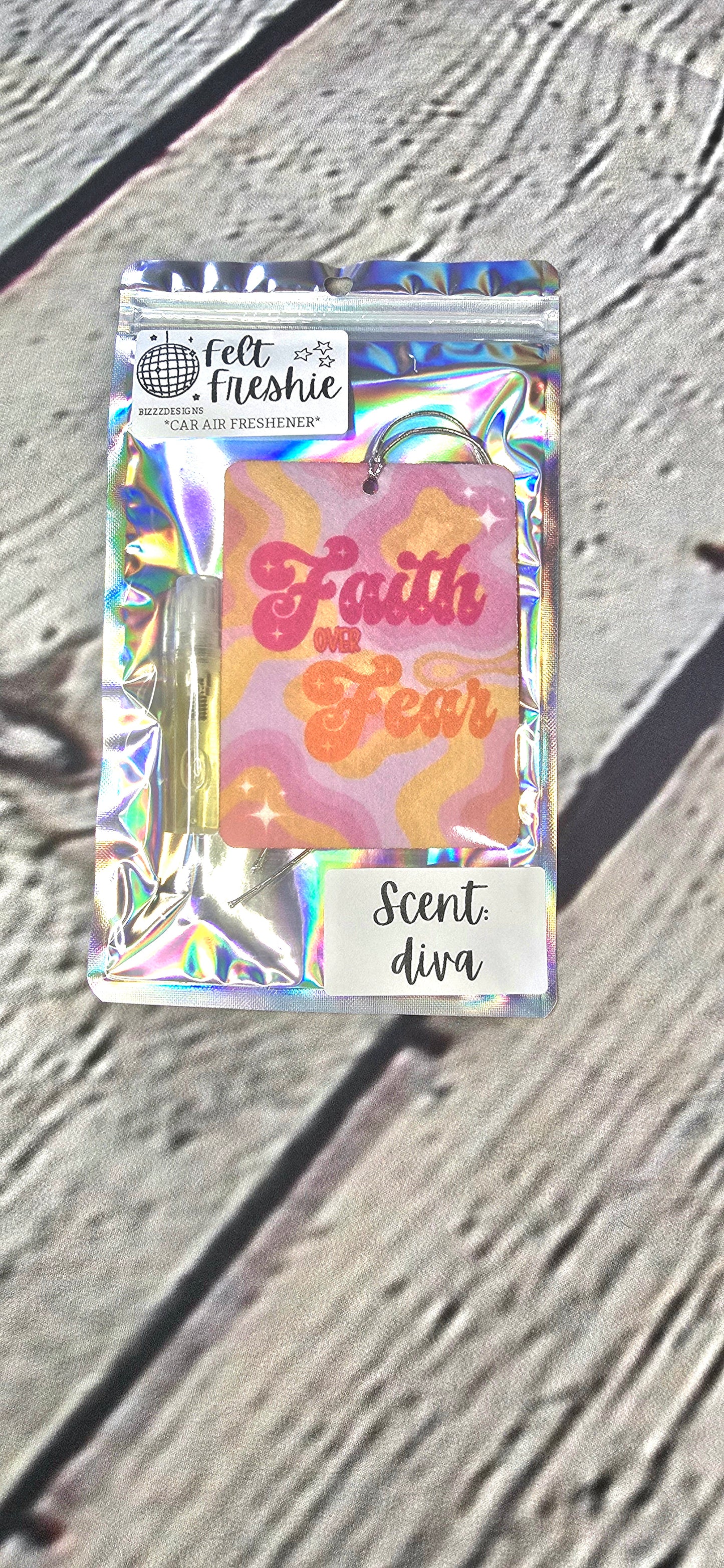 A rectangular felt air freshener with a holoraphic design, featuring the text 'Faith' and the phrase 'Scent diva' on a wooden surface.