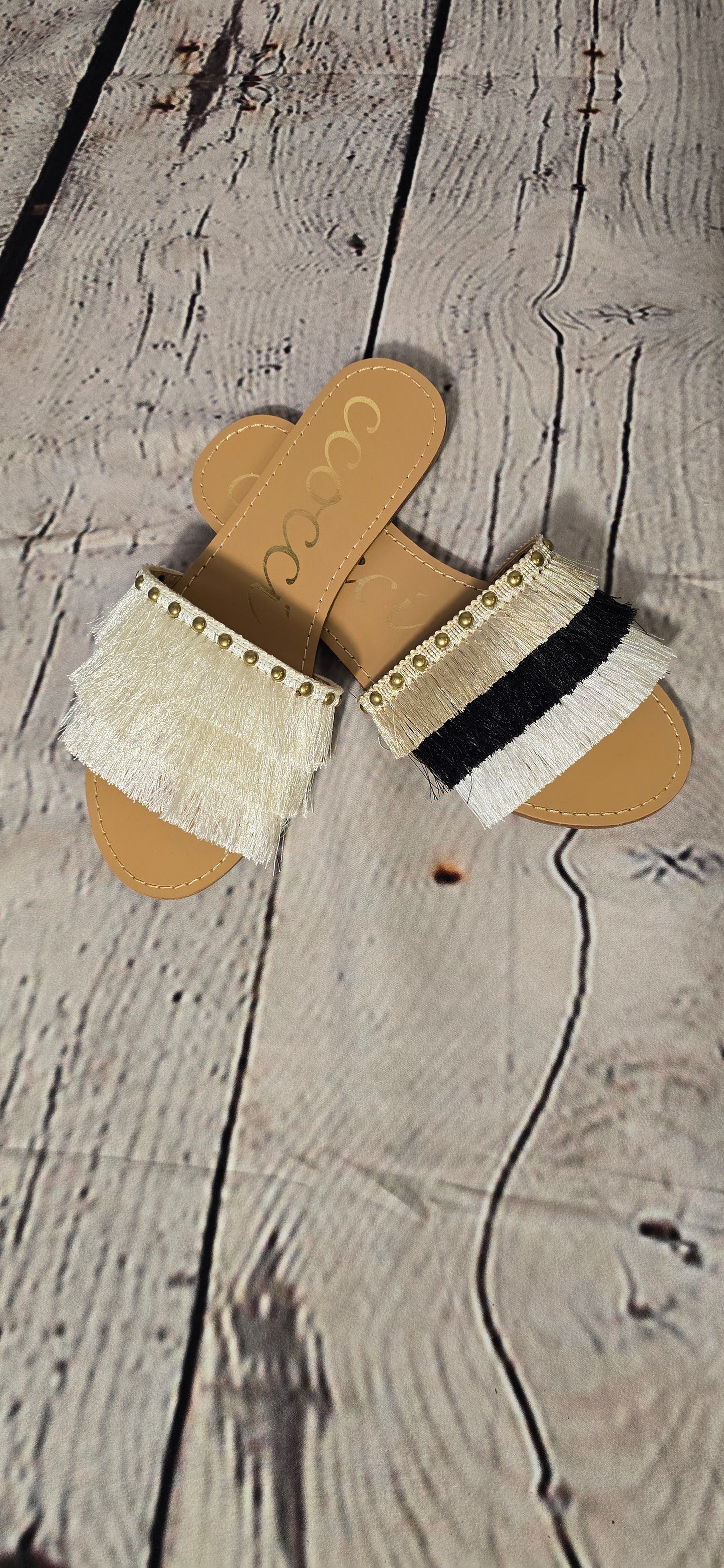 A pair of flat sandals with fine satin fringe and brass stud decorations in beige, black, and white colors.