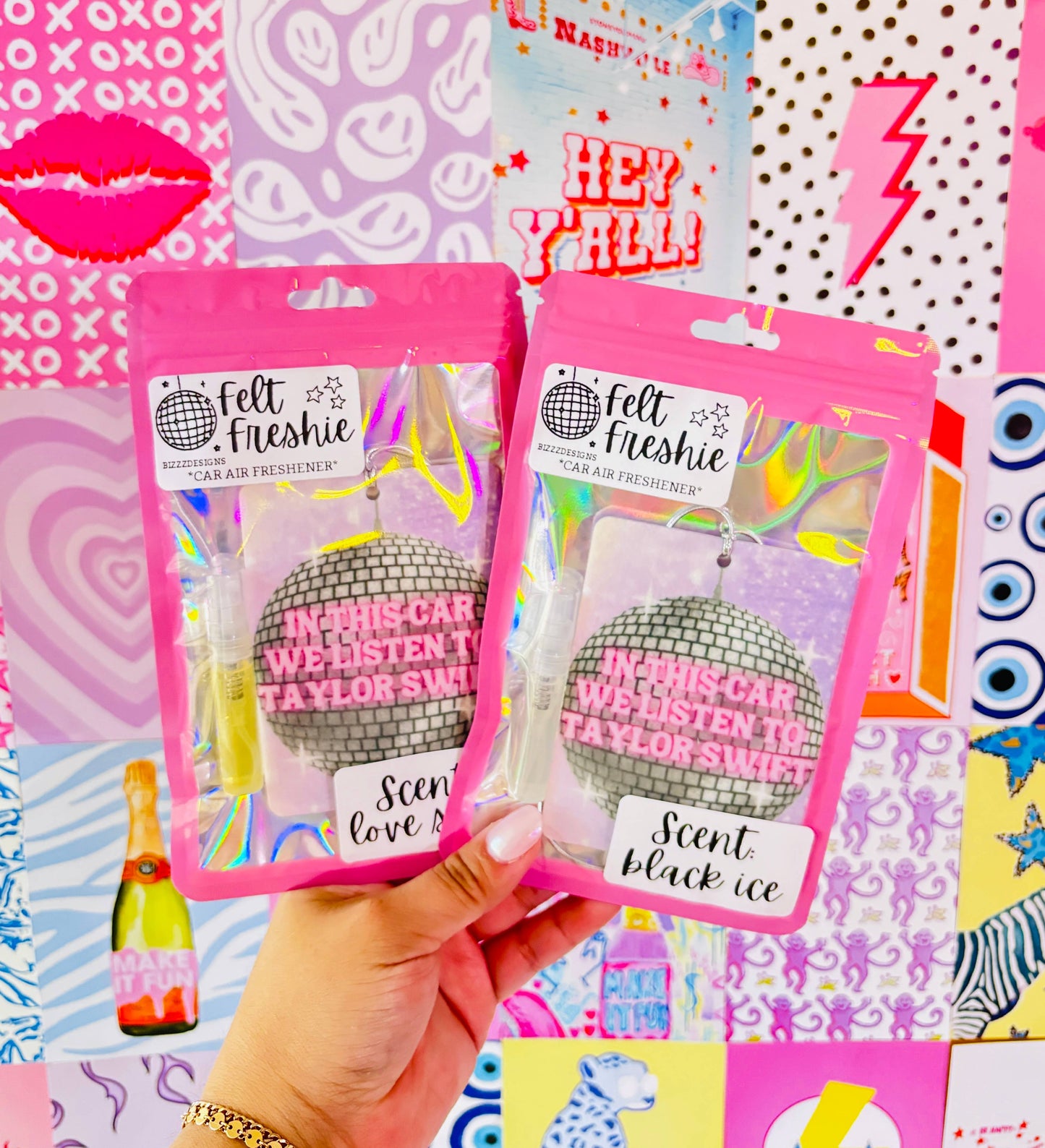 This image features two Swiftie Felt Freshies, designed as car air fresheners with a Taylor Swift theme. The product packaging is vibrant pink, complementing the eye-catching design of the air fresheners that resemble disco balls in silver and blue.