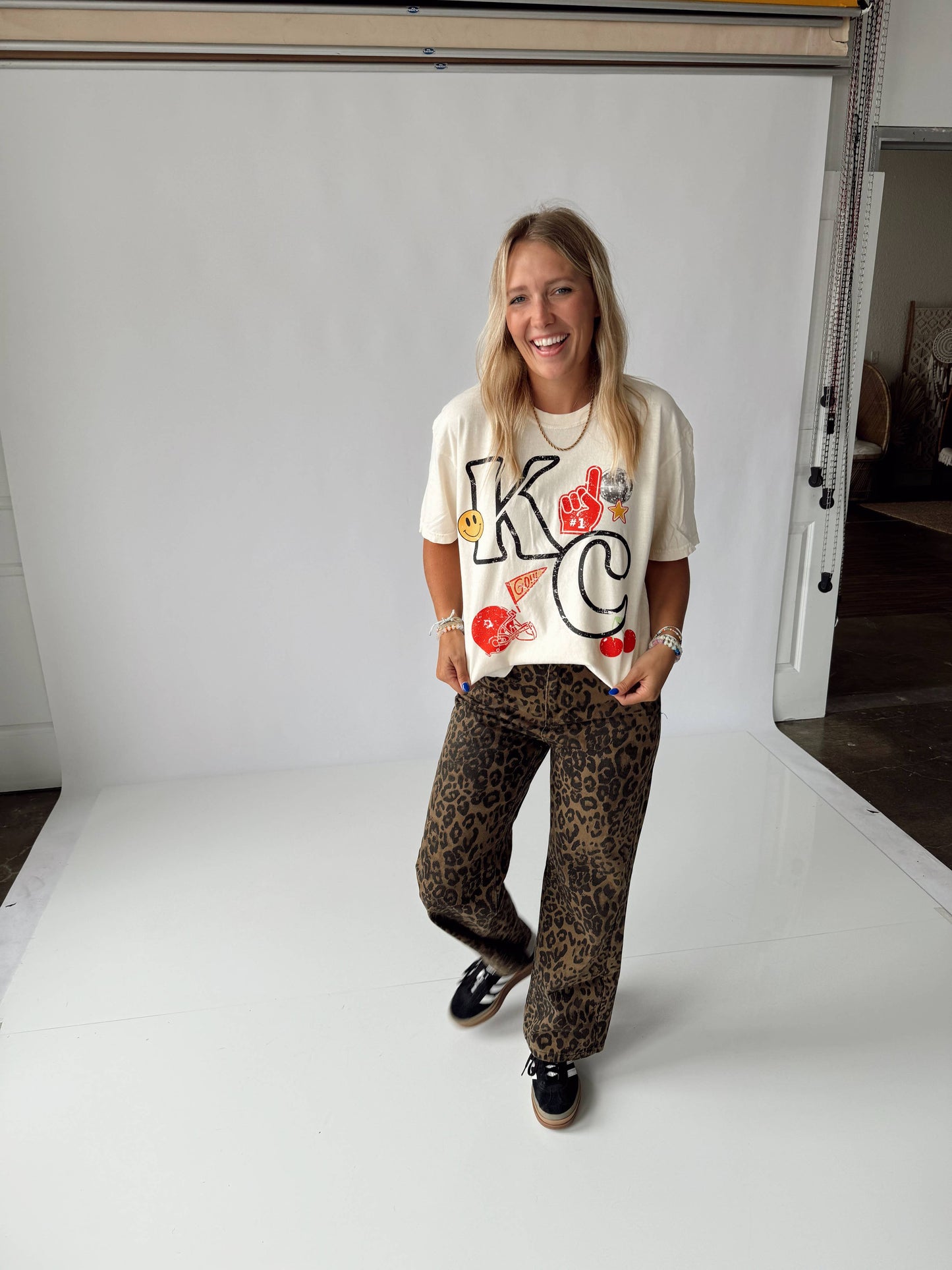 A person wearing a white KC themed t-shirt and animal patterned pants, standing in a room with a white background.