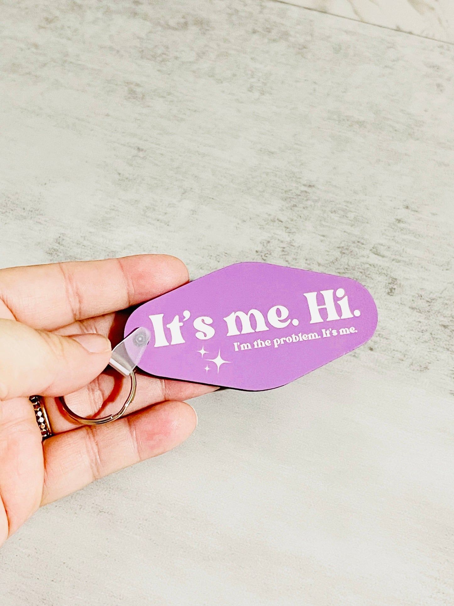 a person holding A double-sided pink keychain with white text that reads 'It's me. Hi. I'm the problem. It's me.' on a textured background.