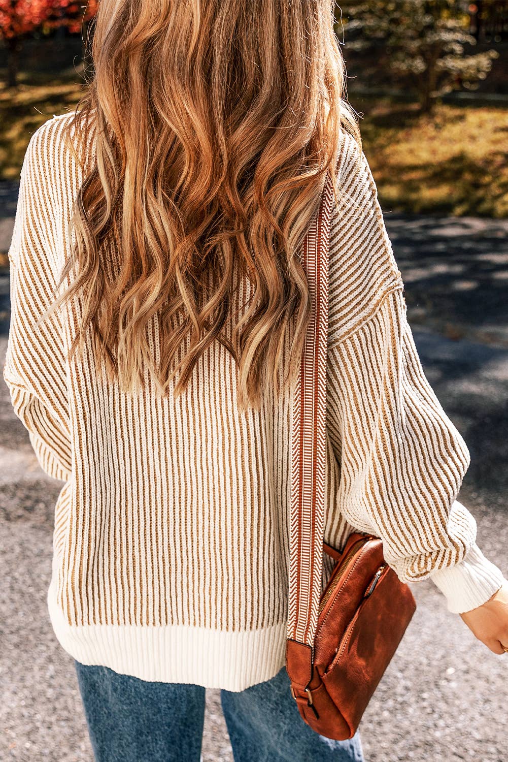 A woman wearing a striped contrast trim loose sweater in beige and white, paired with blue jeans and a brown bag.