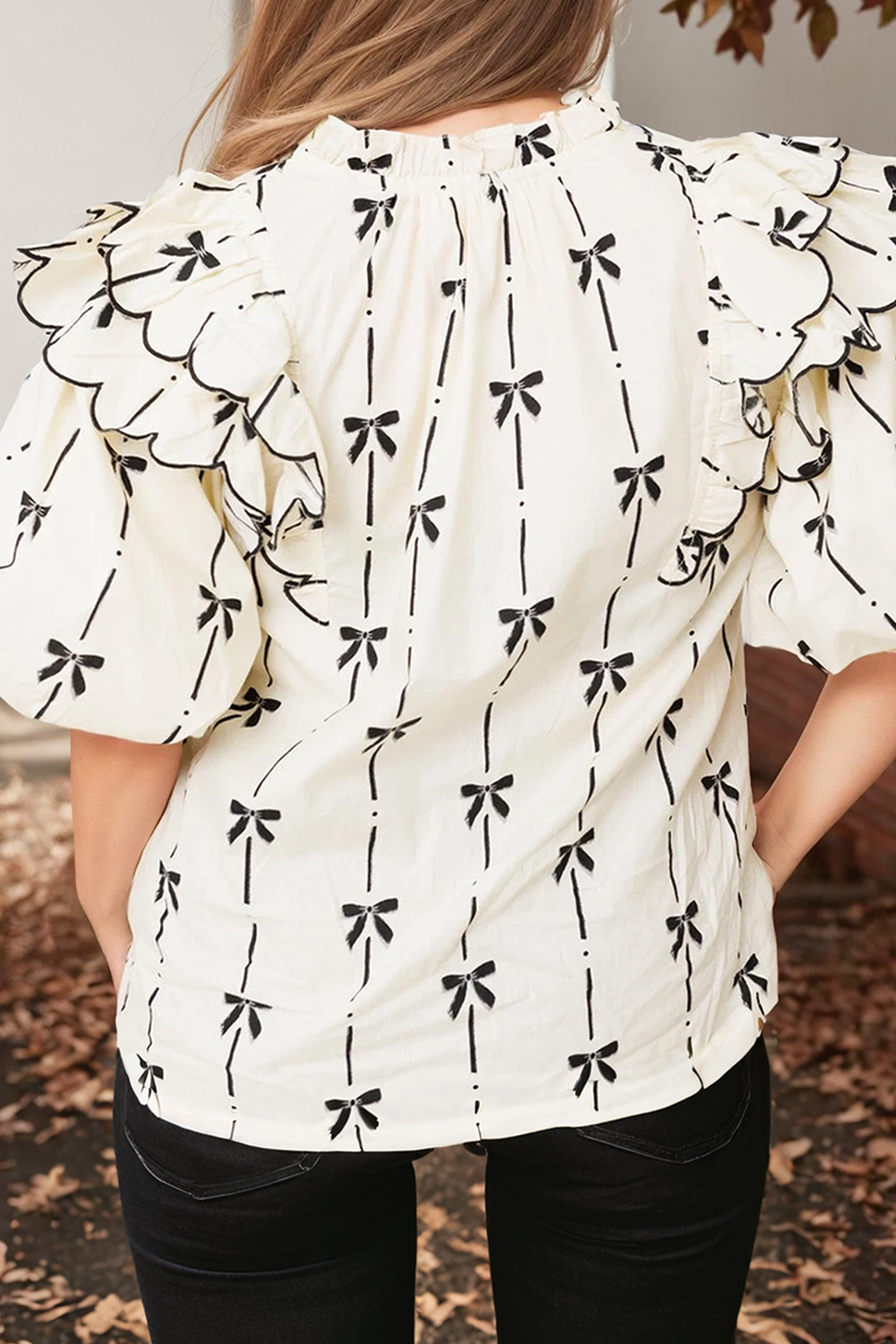 A woman standing against a brick wall background, wearing a white ruffled bubble sleeve notched neck blouse with a black pattern.