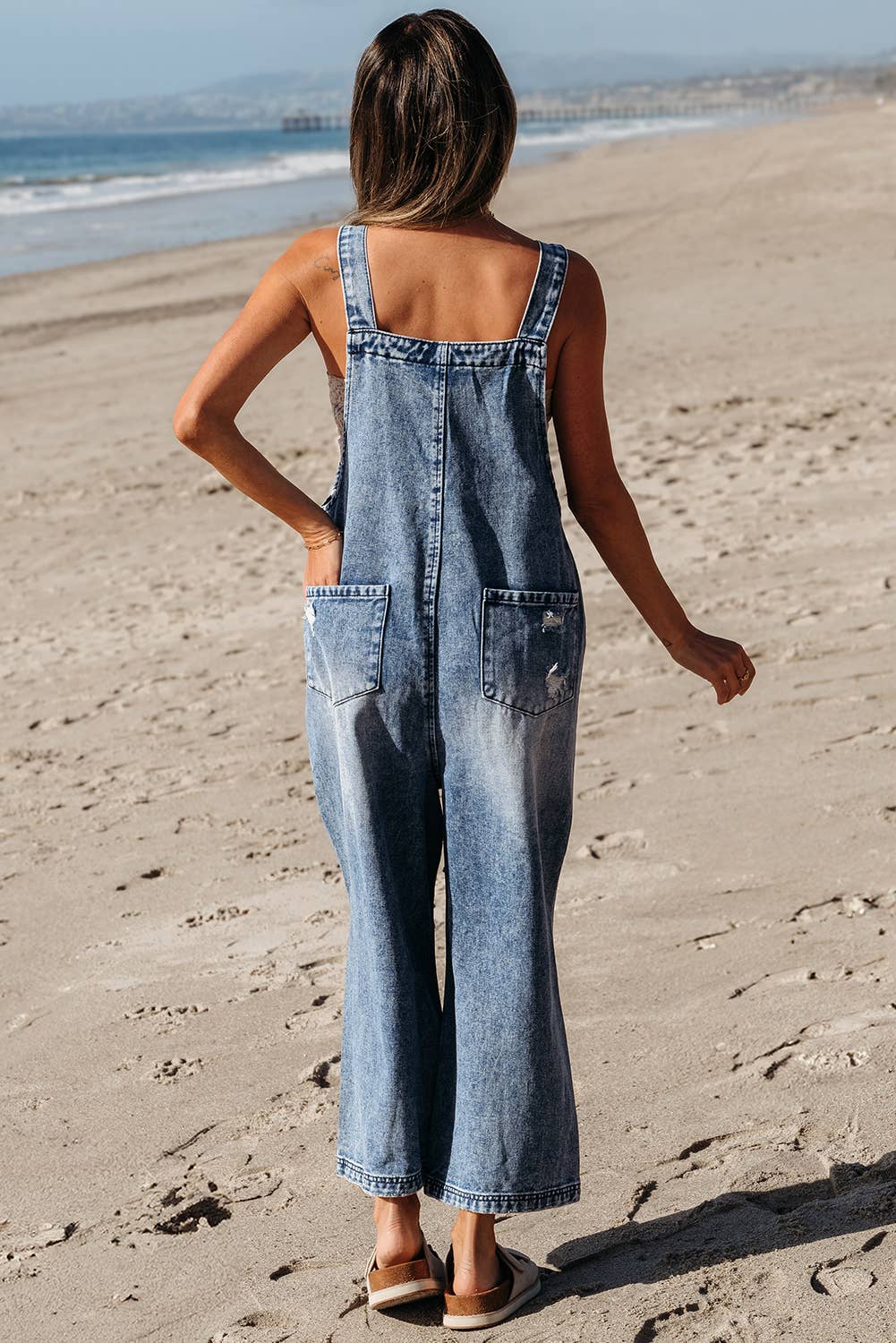 Person wearing denim overalls standing on a sandy beach.