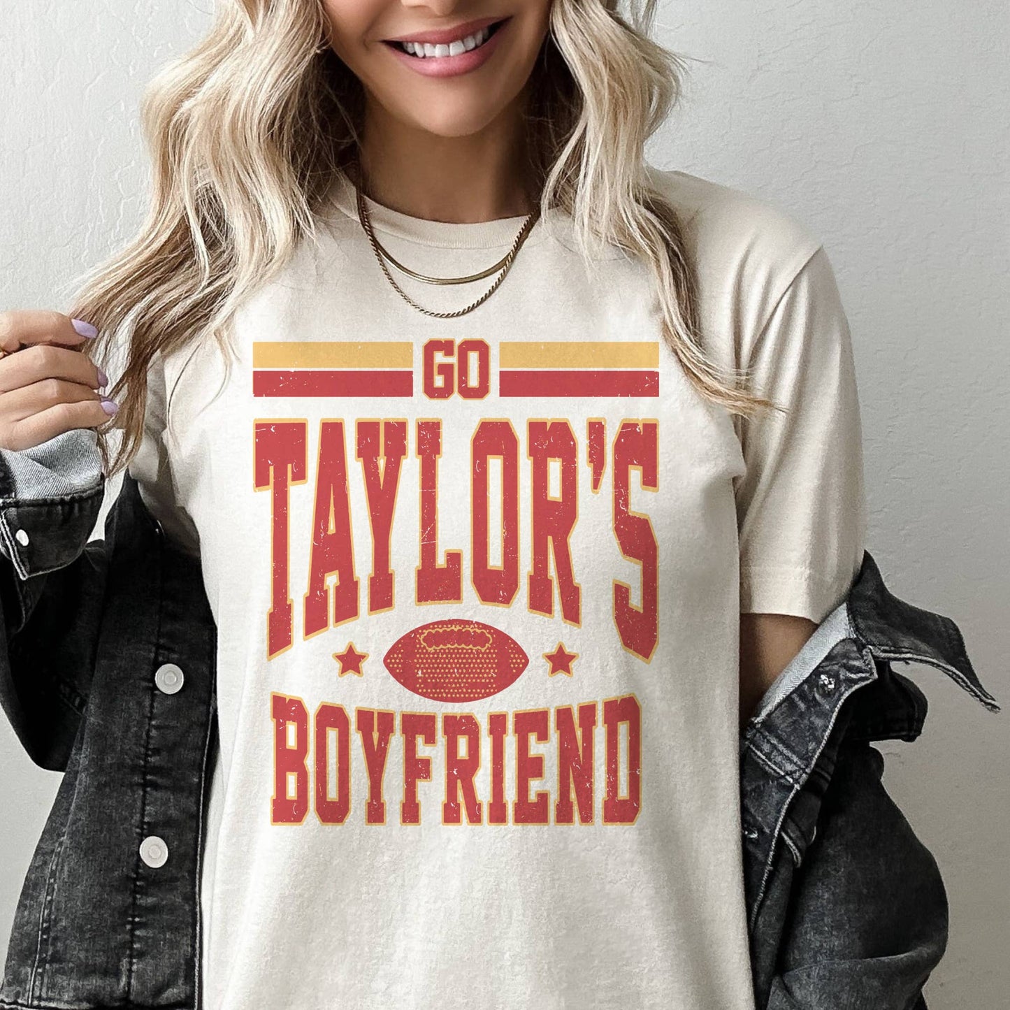 A person wearing a beige graphic tee with the text 'GO TAYLOR'S BOYFRIEND' followed by 'Kansas City Football' in a stylized font, predominantly in red and yellow colors.