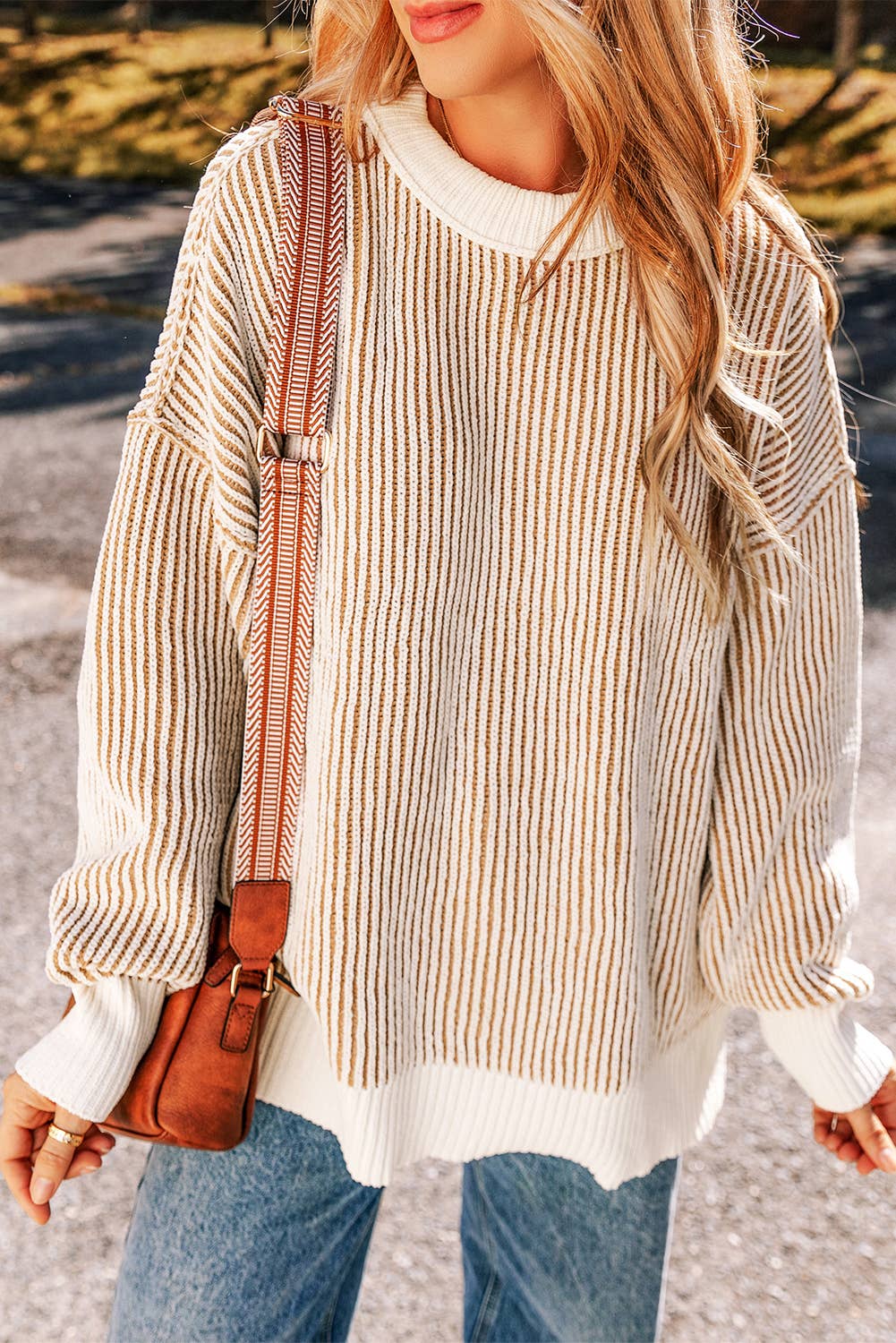 A woman wearing a striped contrast trim loose sweater in beige and white, paired with blue jeans and a brown bag.