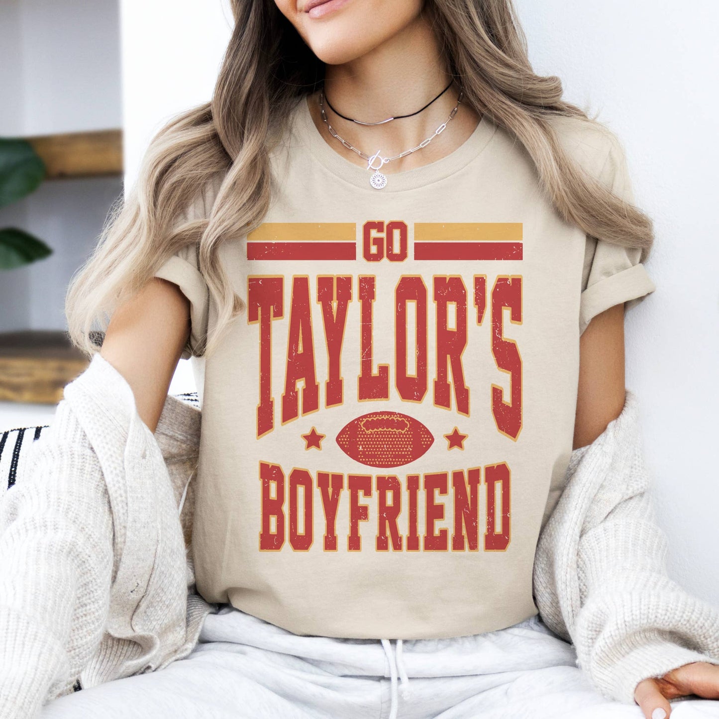 A person wearing a beige graphic tee with the text 'GO TAYLOR'S BOYFRIEND' followed by 'Kansas City Football' in a stylized font, predominantly in red and yellow colors.