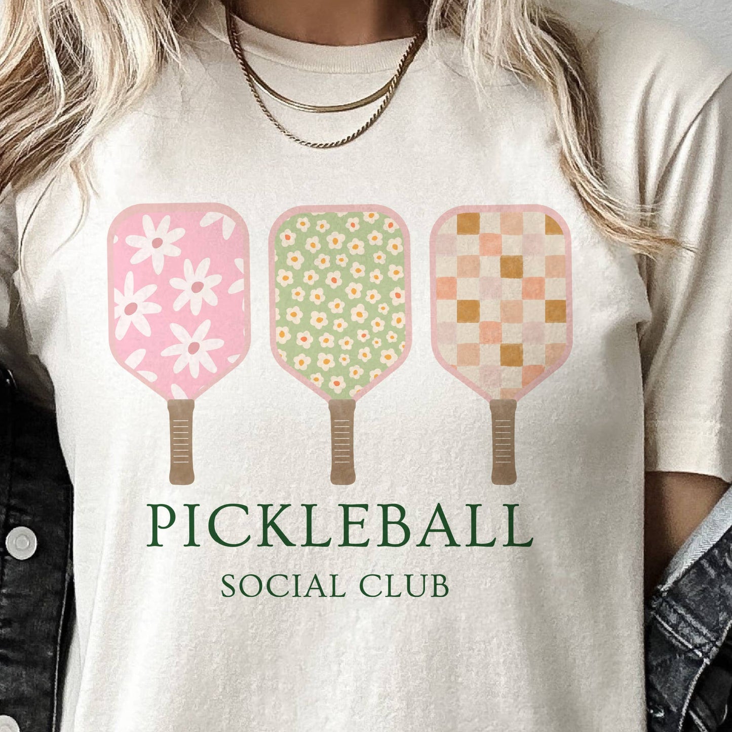 A person wearing a white t-shirt with 'PICKLEBALL SOCIAL CLUB' text and a graphic of three pickleball paddles in pink, green, and yellow colors with floral and checkered patterns.