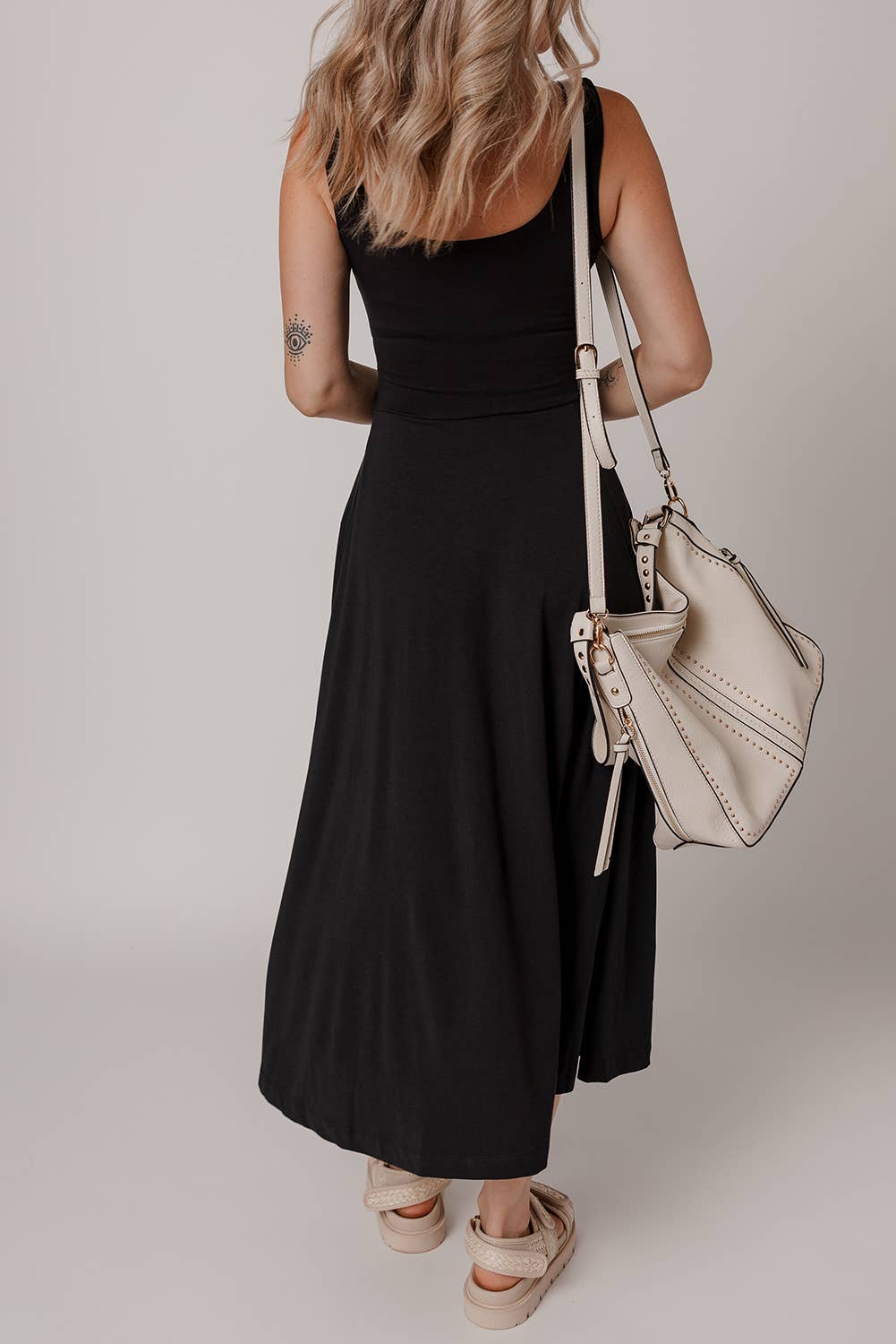A woman wearing a black solid side split midi dress with no visible sleeves, paired with a white bag and beige footwear. She has accessorized with a necklace.