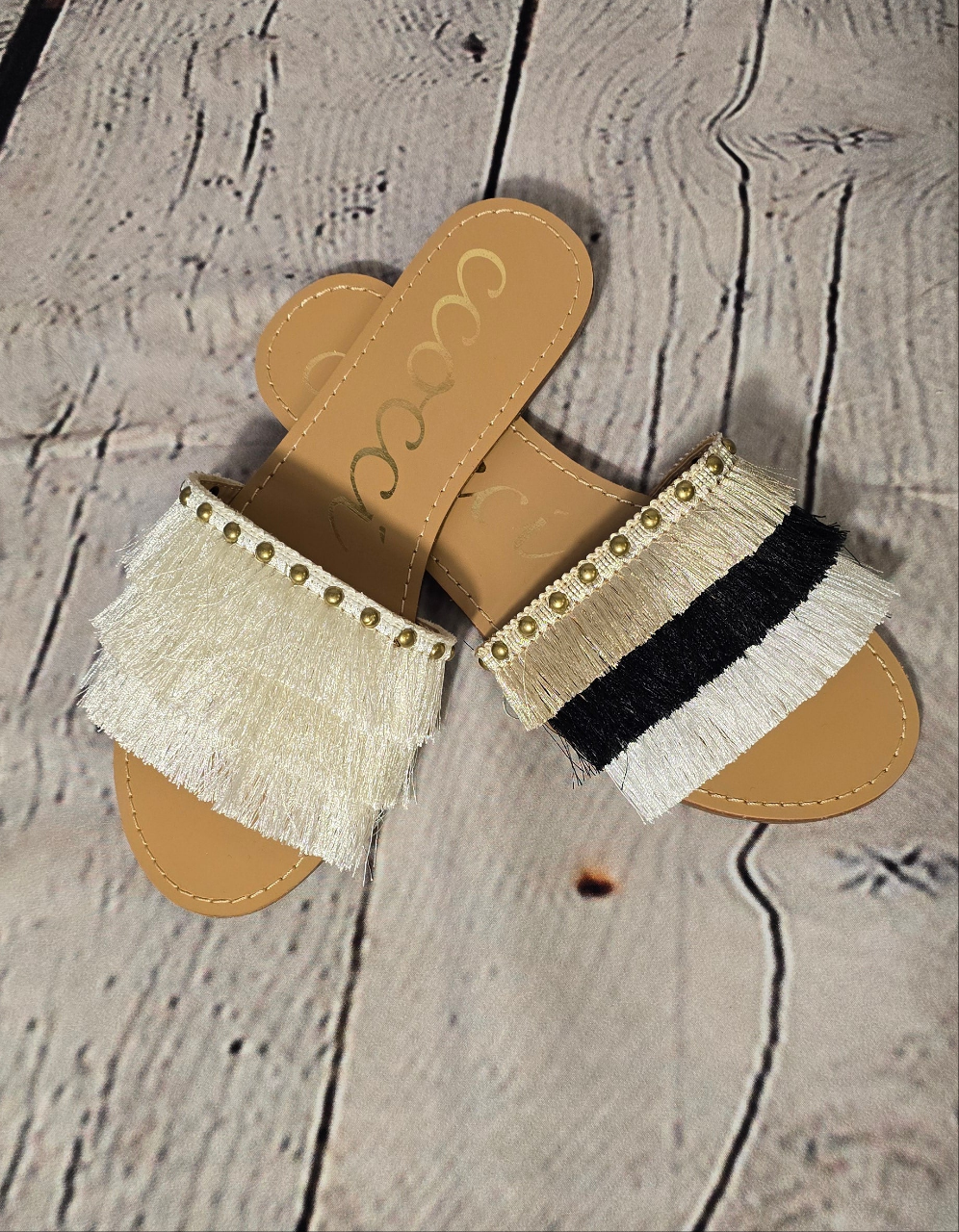 A pair of flat sandals with fine satin fringe and brass stud decorations in beige, black, and white colors.