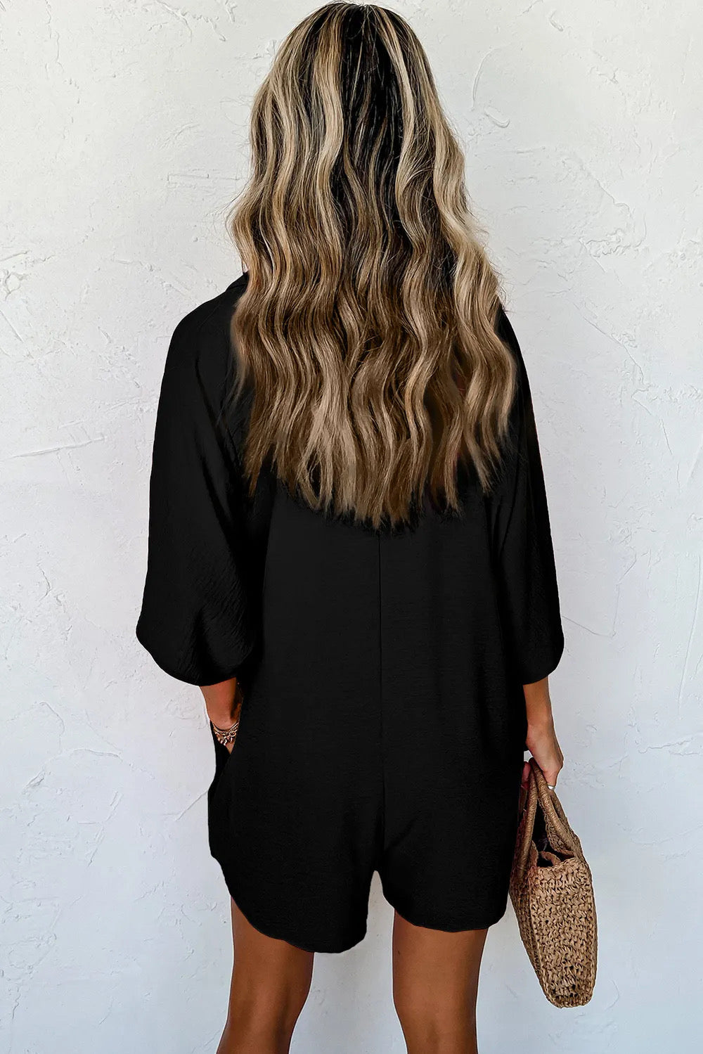 A person wearing a black short-sleeved collared romper with half buttons, paired with a wide-brimmed hat and gold accessories.