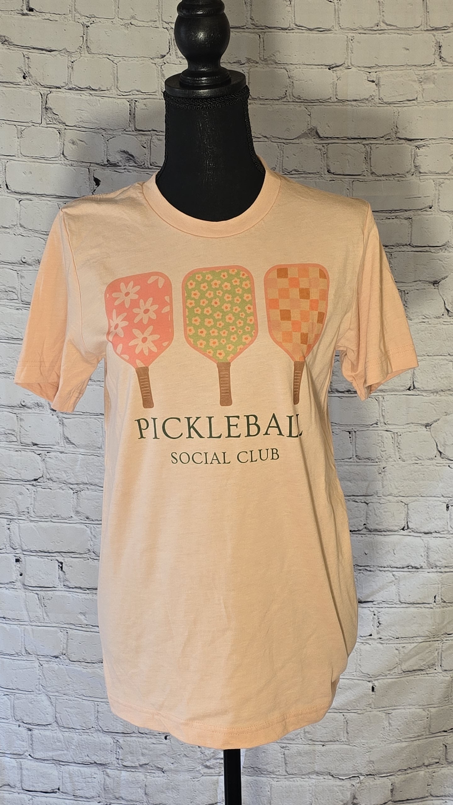 peach t-shirt with 'PICKLEBALL SOCIAL CLUB' text and a graphic of three pickleball paddles in pink, green, and yellow colors with floral and checkered patterns.