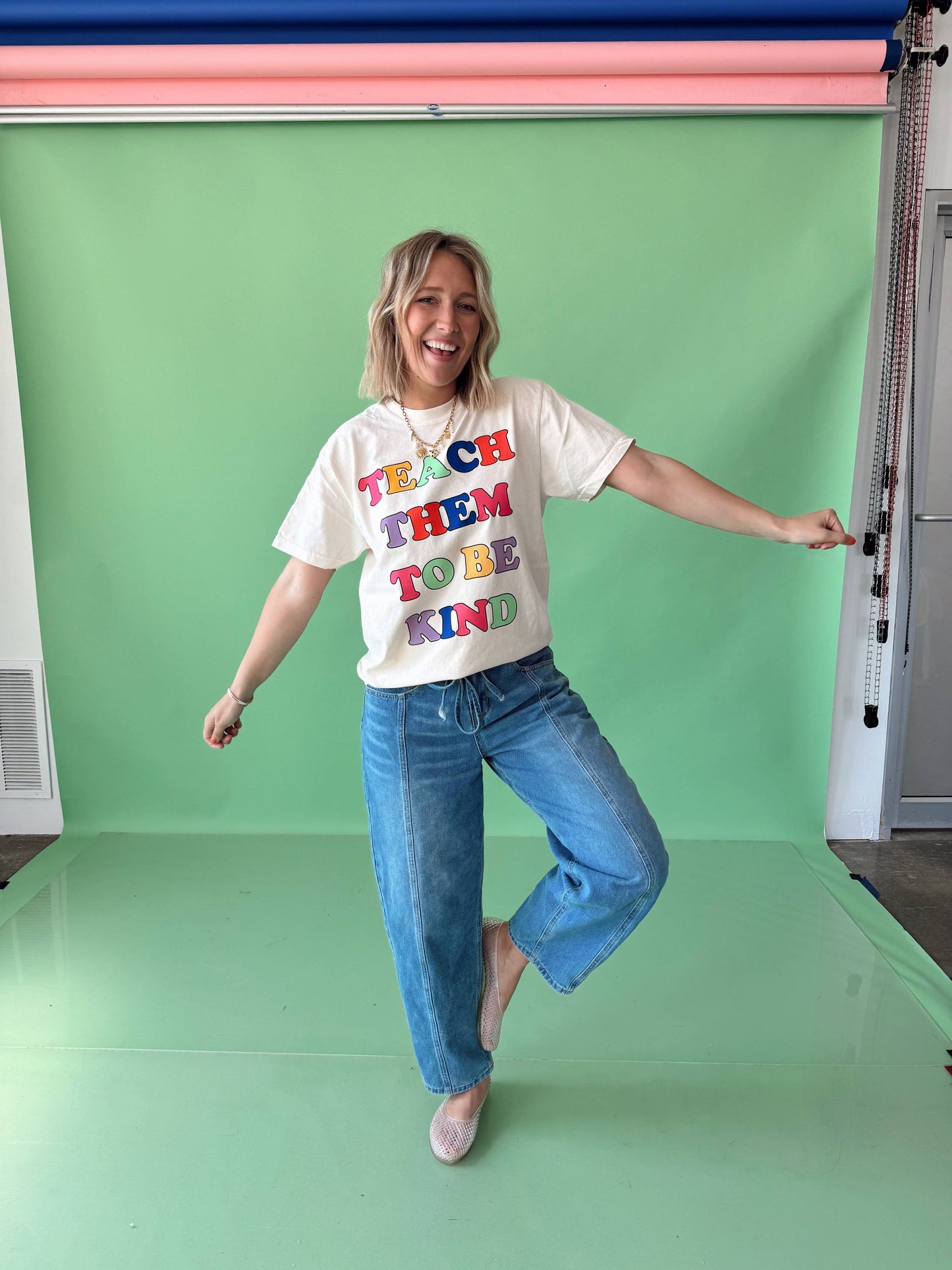 A person wearing a white t-shirt with the text 'TEACH THEM TO BE KIND' in colorful letters, paired with blue jeans and beige shoes.
