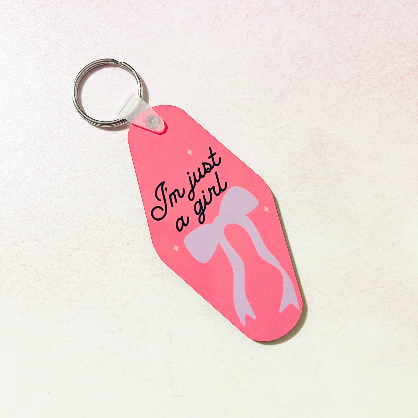 A pink keychain with the text 'I'm just a girl' printed on it, featuring a bow design.