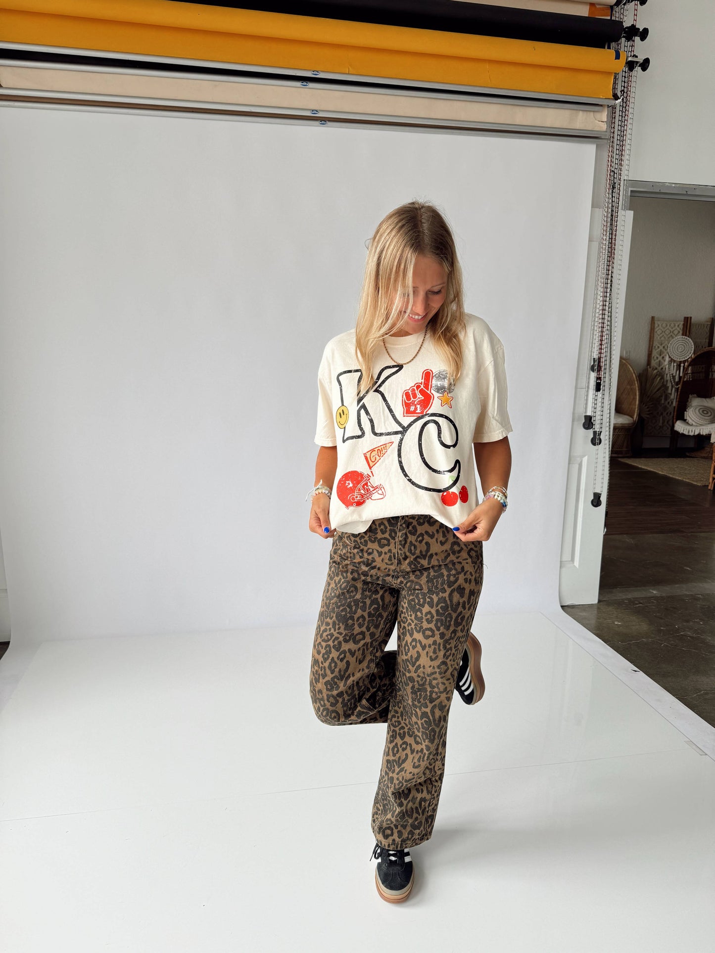 A person wearing a white KC themed t-shirt and animal patterned pants, standing in a room with a white background.
