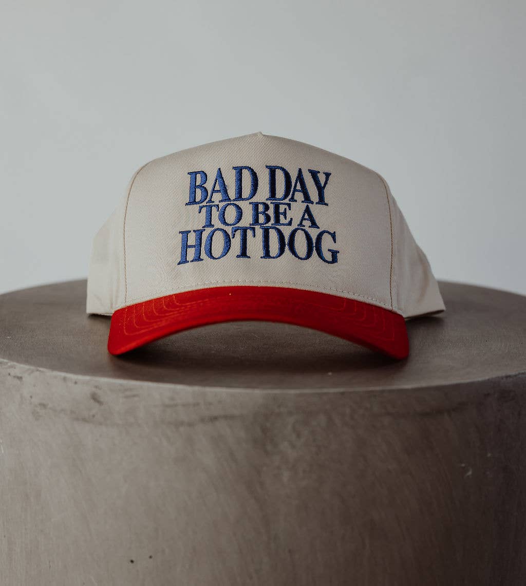 Beige cap with red brim and blue text 'Bad Day to be a Hotdog' on a wooden surface.