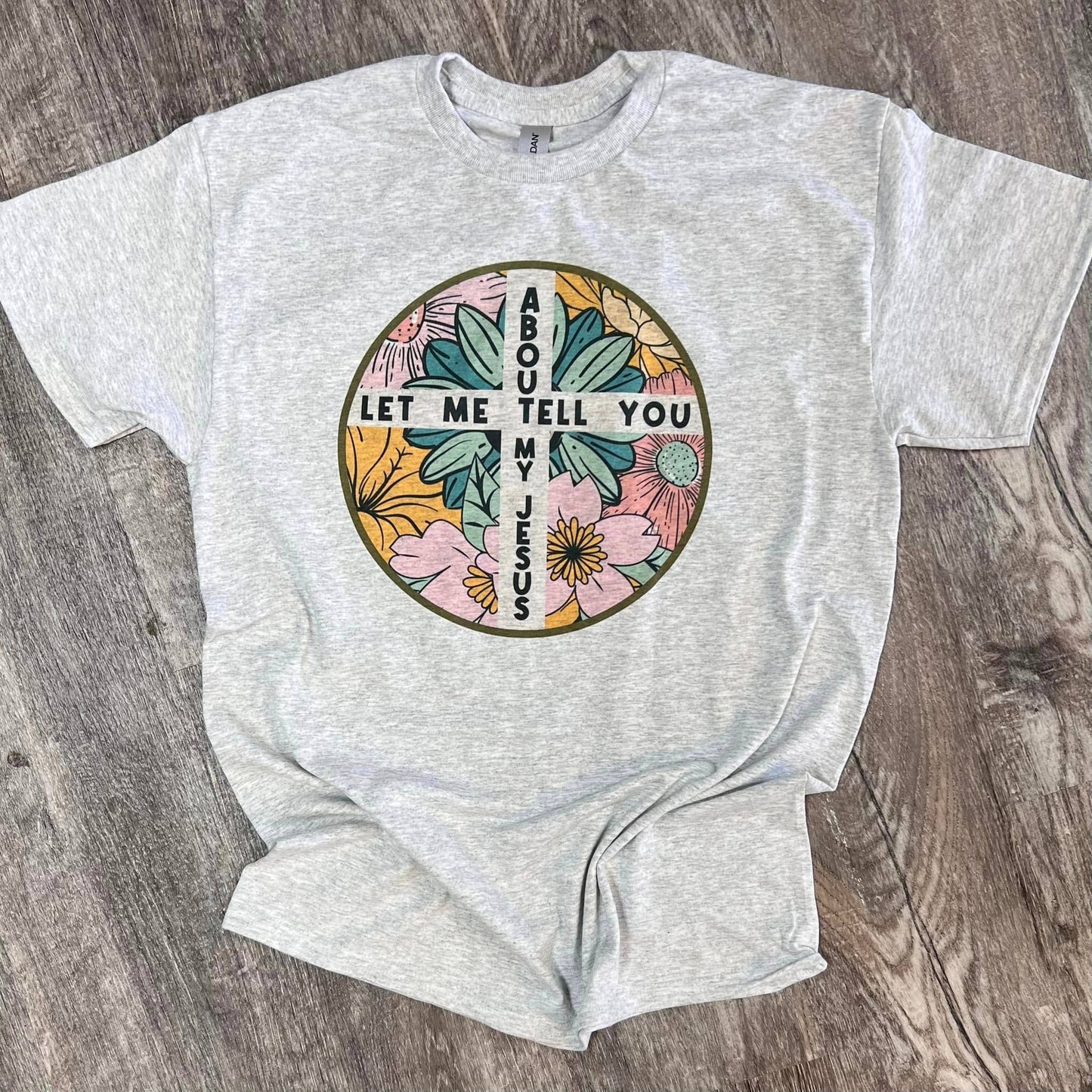 A gray unisex t-shirt with a round floral design containing text that reads 'LET ME TELL YOU ABOUT MY JESUS'.