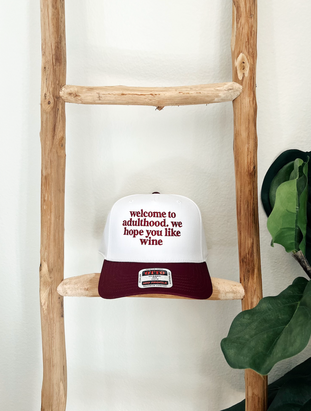 A maroon and white baseball cap with the text 'Welcome to adulthood.