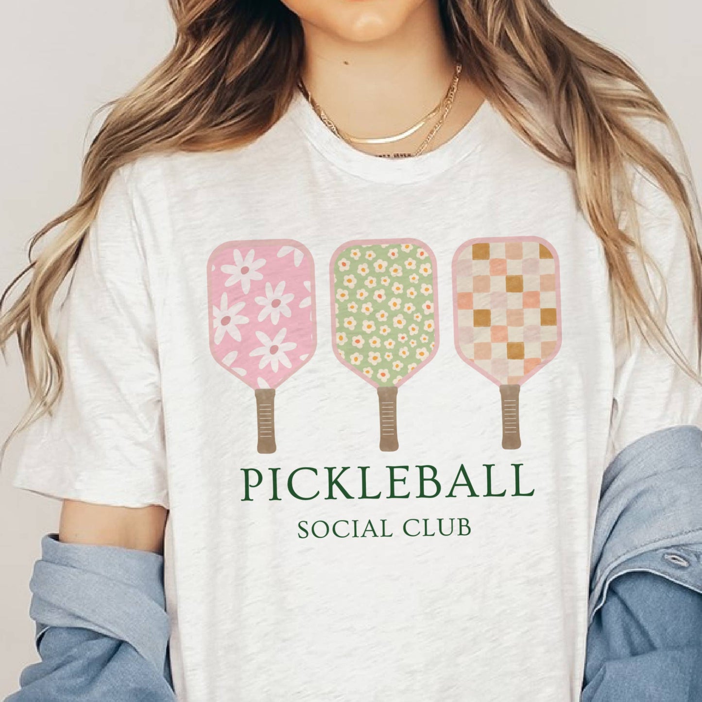 A person wearing a white t-shirt with 'PICKLEBALL SOCIAL CLUB' text and a graphic of three pickleball paddles in pink, green, and yellow colors with floral and checkered patterns.