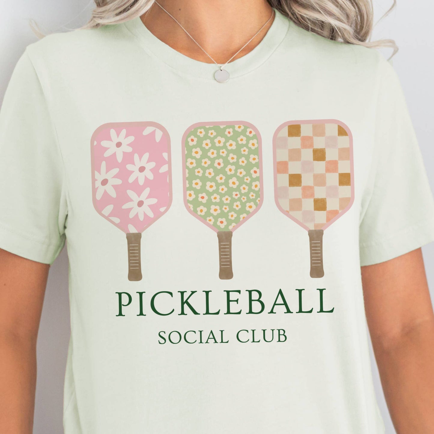 A person wearing a white t-shirt with 'PICKLEBALL SOCIAL CLUB' text and a graphic of three pickleball paddles in pink, green, and yellow colors with floral and checkered patterns.