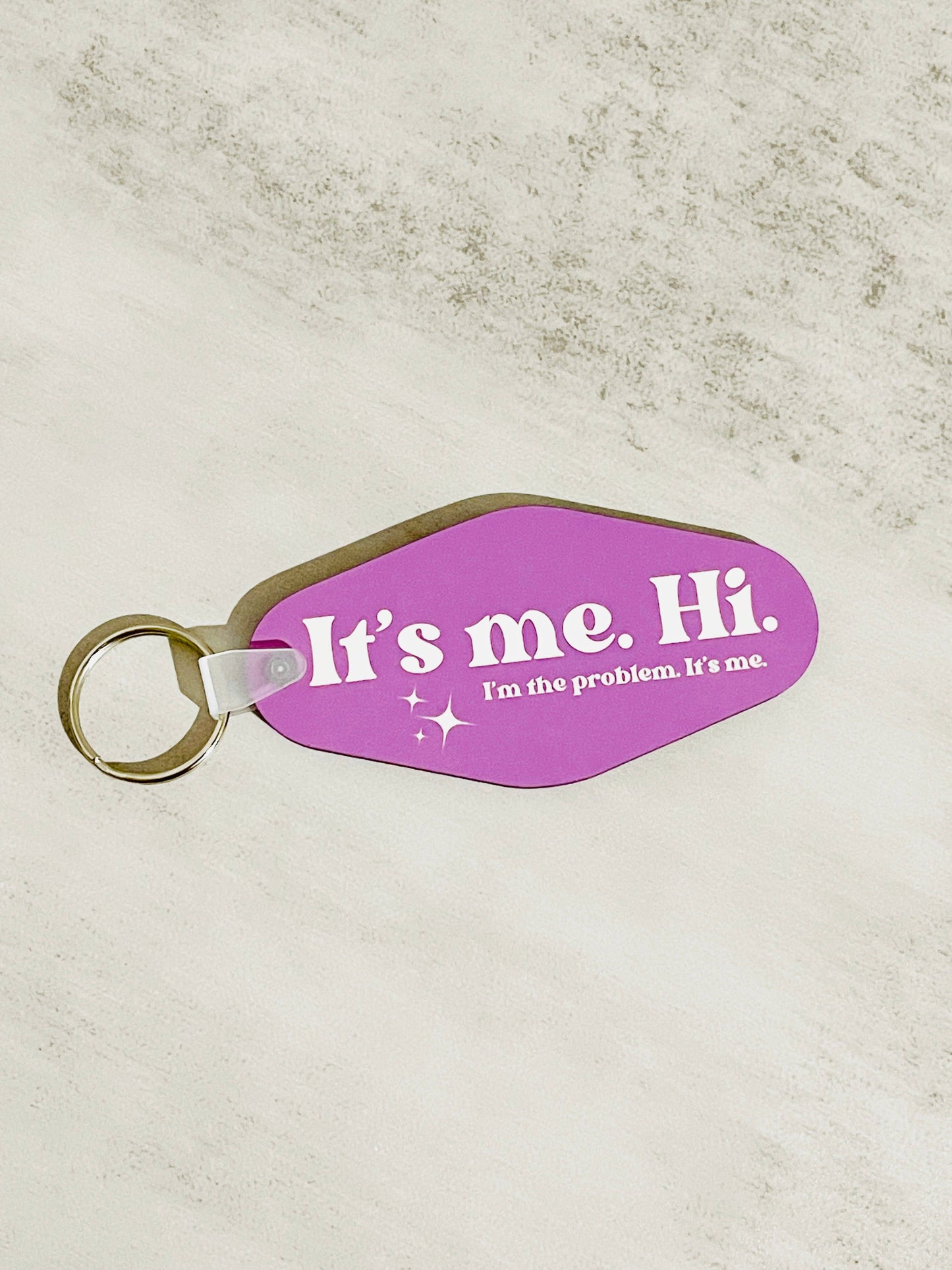 A double-sided pink keychain with white text that reads 'It's me. Hi. I'm the problem. It's me.' on a textured background.