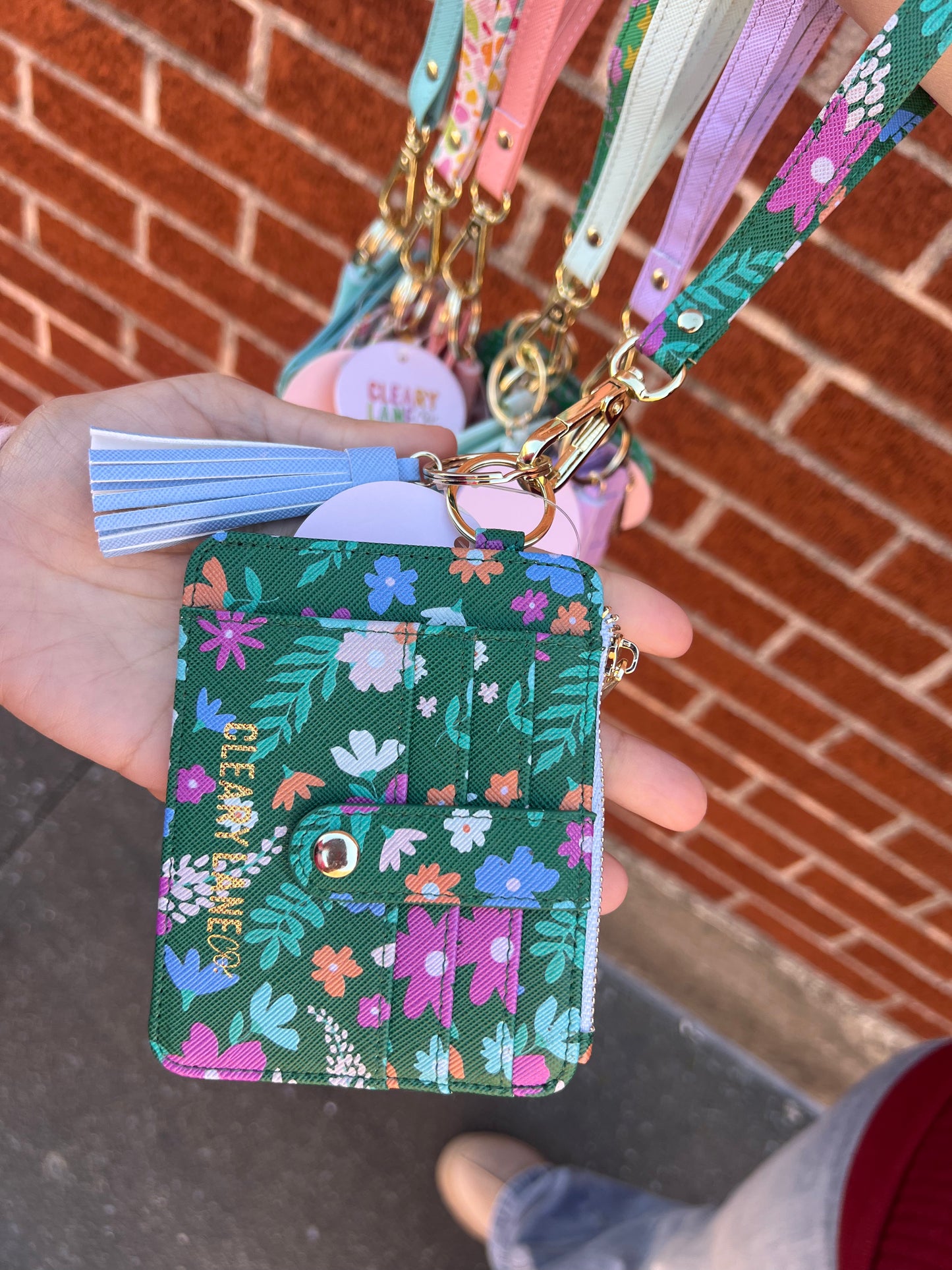A hand holding a green wallet keychain with a floral pattern and gold hardware. The keychain is attached to a strap and there are multiple keys on it.