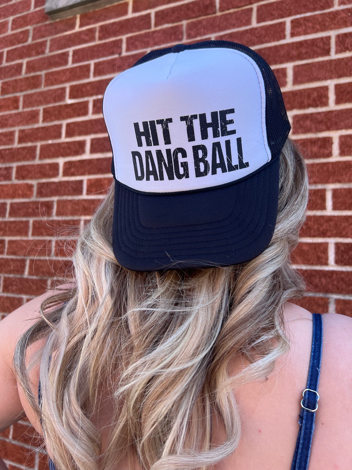 A person wearing a black and white foam trucker hat with the text 'HIT THE DANG BALL' printed on the front.