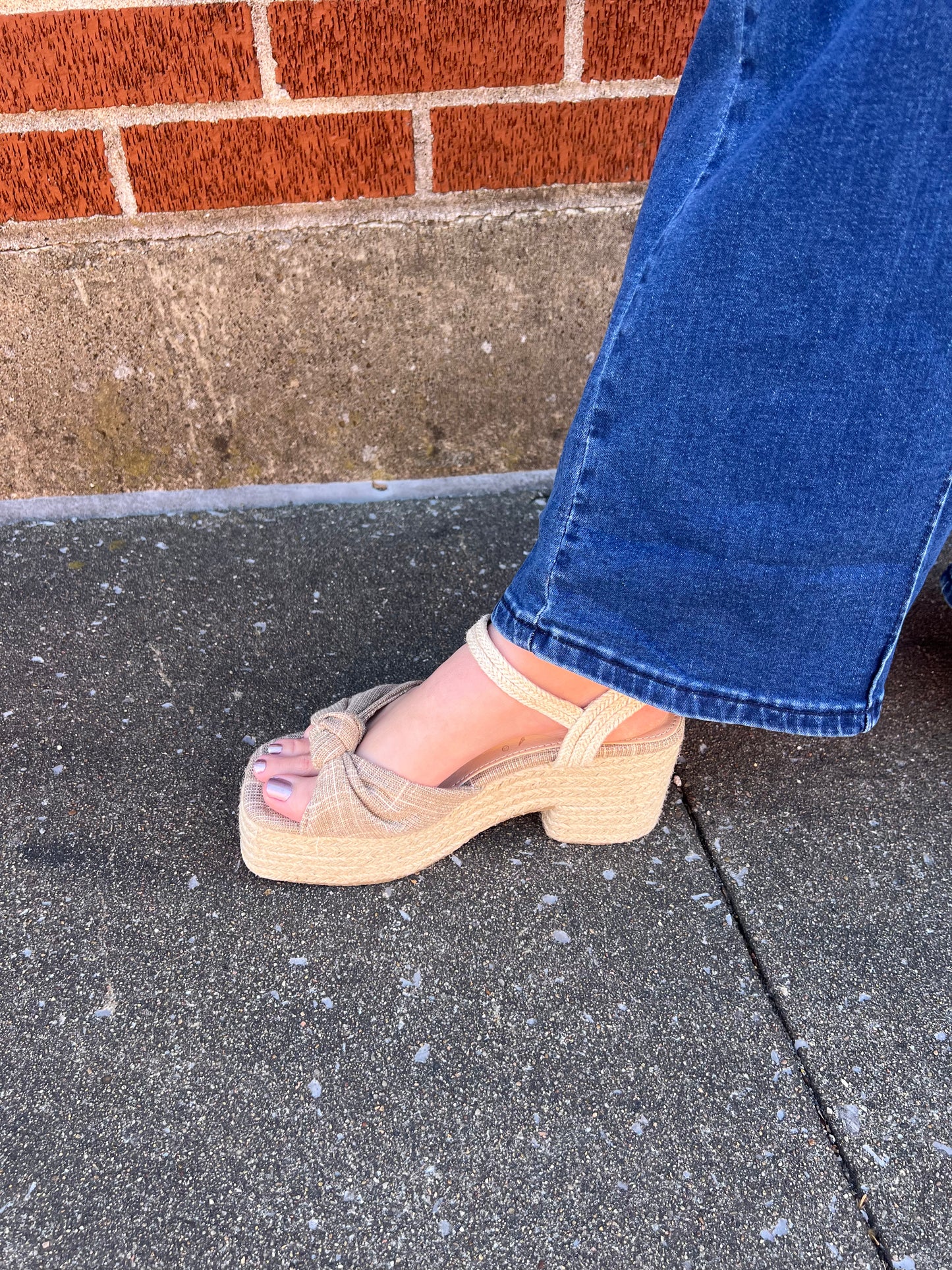 A person wearing blue jeans and a beige knotted espadrille platform sandal with a braided raffia ankle strap and gold hardware.