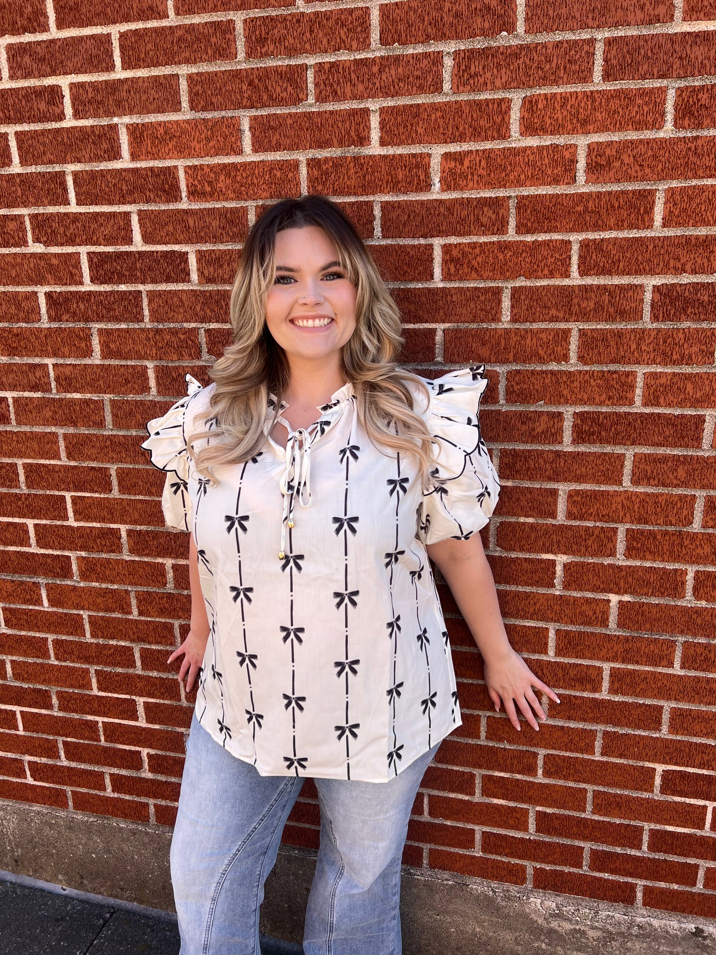 A woman standing against a brick wall background, wearing a white ruffled bubble sleeve notched neck blouse with a black pattern.