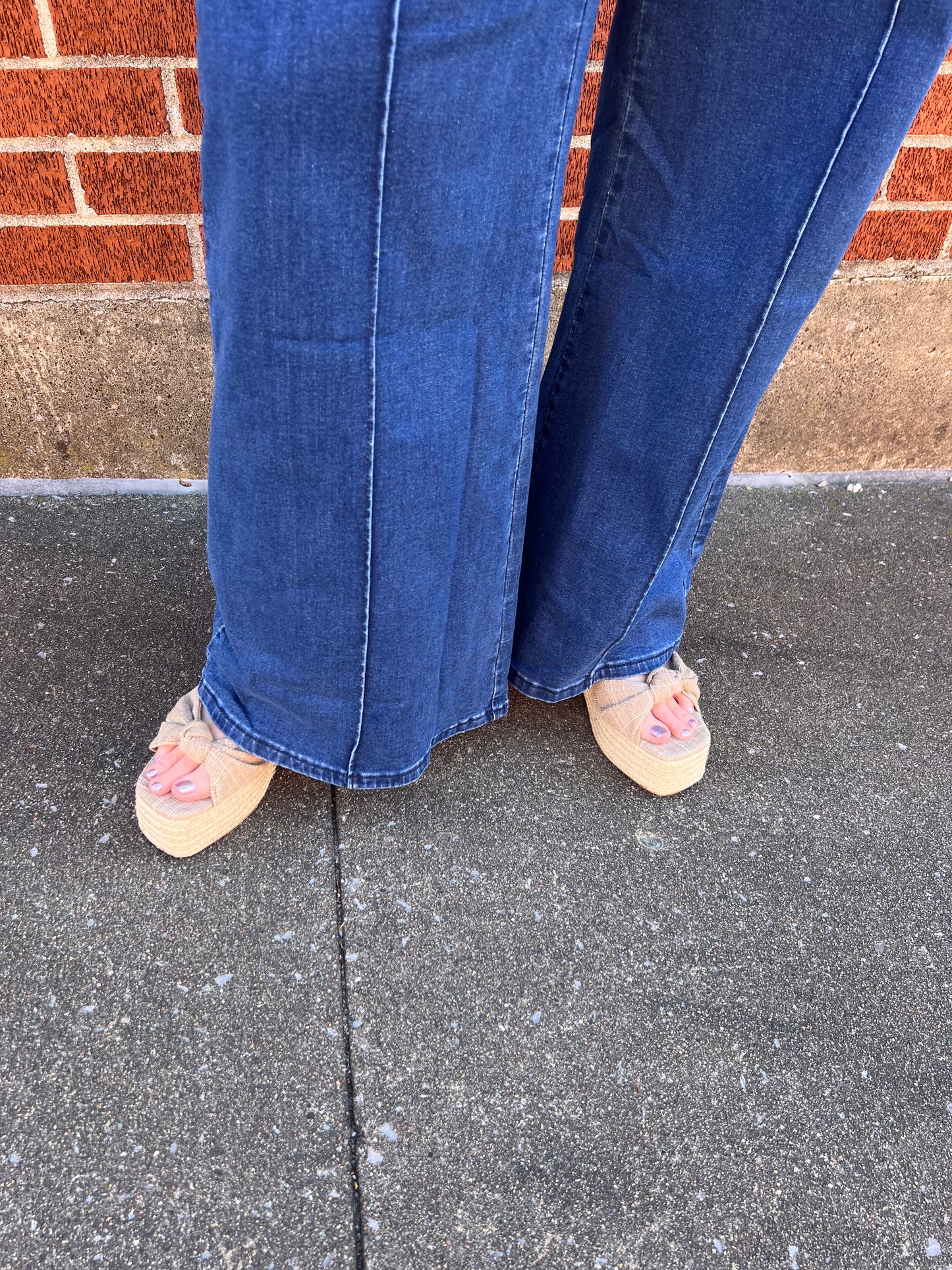 A person wearing blue jeans and a beige knotted espadrille platform sandal with a braided raffia ankle strap and gold hardware.