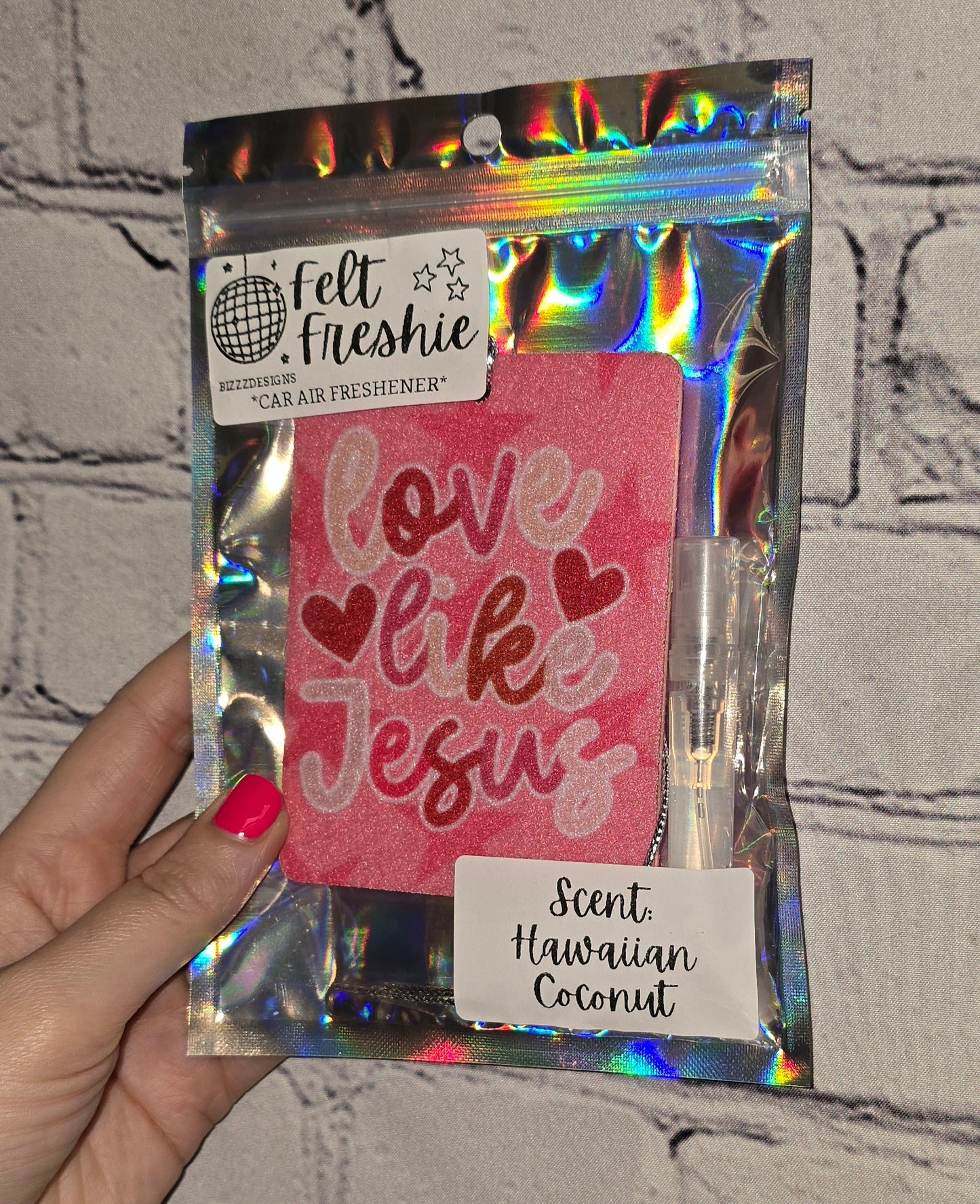 A hand holding a car freshener pack with 'Love Like Jesus' text and heart shapes, accompanied by a 3ml spray bottle.
