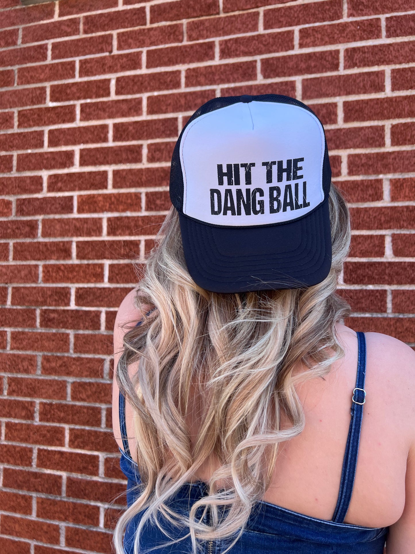 A person wearing a black and white foam trucker hat with the text 'HIT THE DANG BALL' printed on the front.