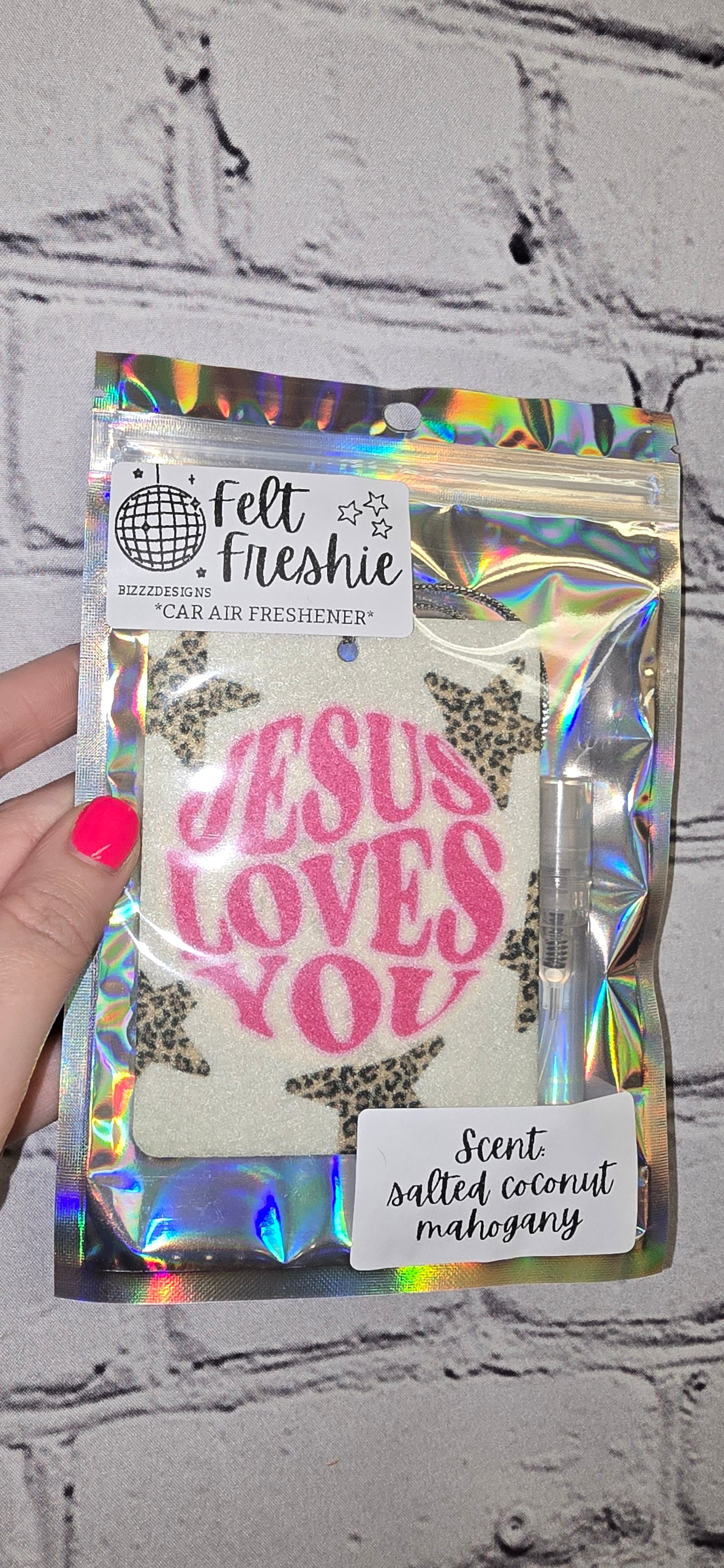 A hand holding a car freshener pack with 'JESUS LOVES YOU' text visible on the front. The pack is predominantly white with a pattern and the text is in pink.
