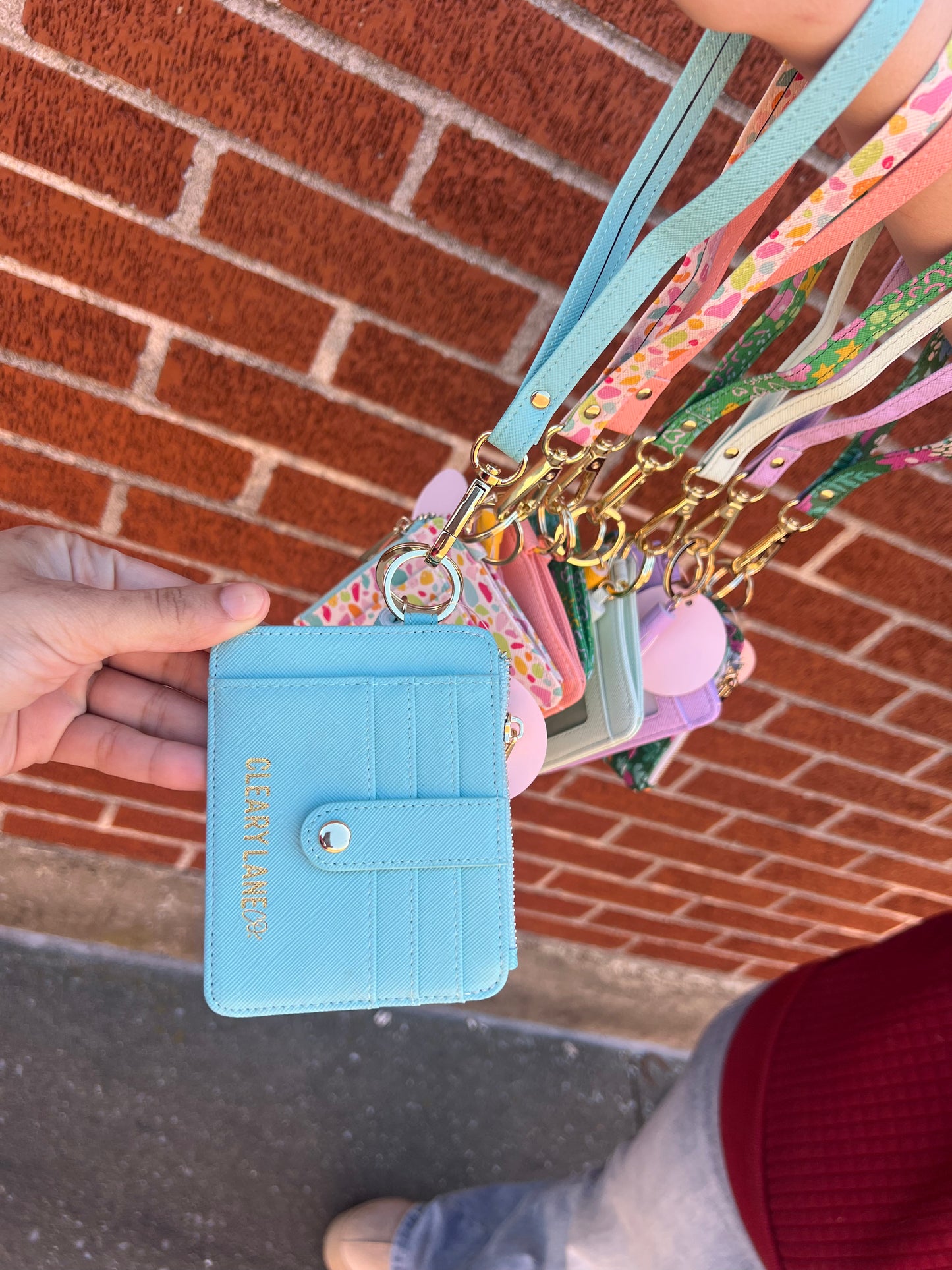 A person holding out a blue wallet keychain with multiple colorful keychains attached to it, displayed against a brick wall.