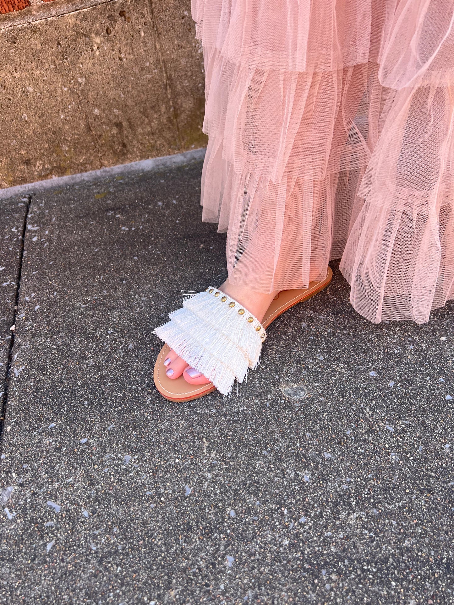 A pair of flat sandals with fringe detailing on the straps, worn by a person standing on a textured ground.