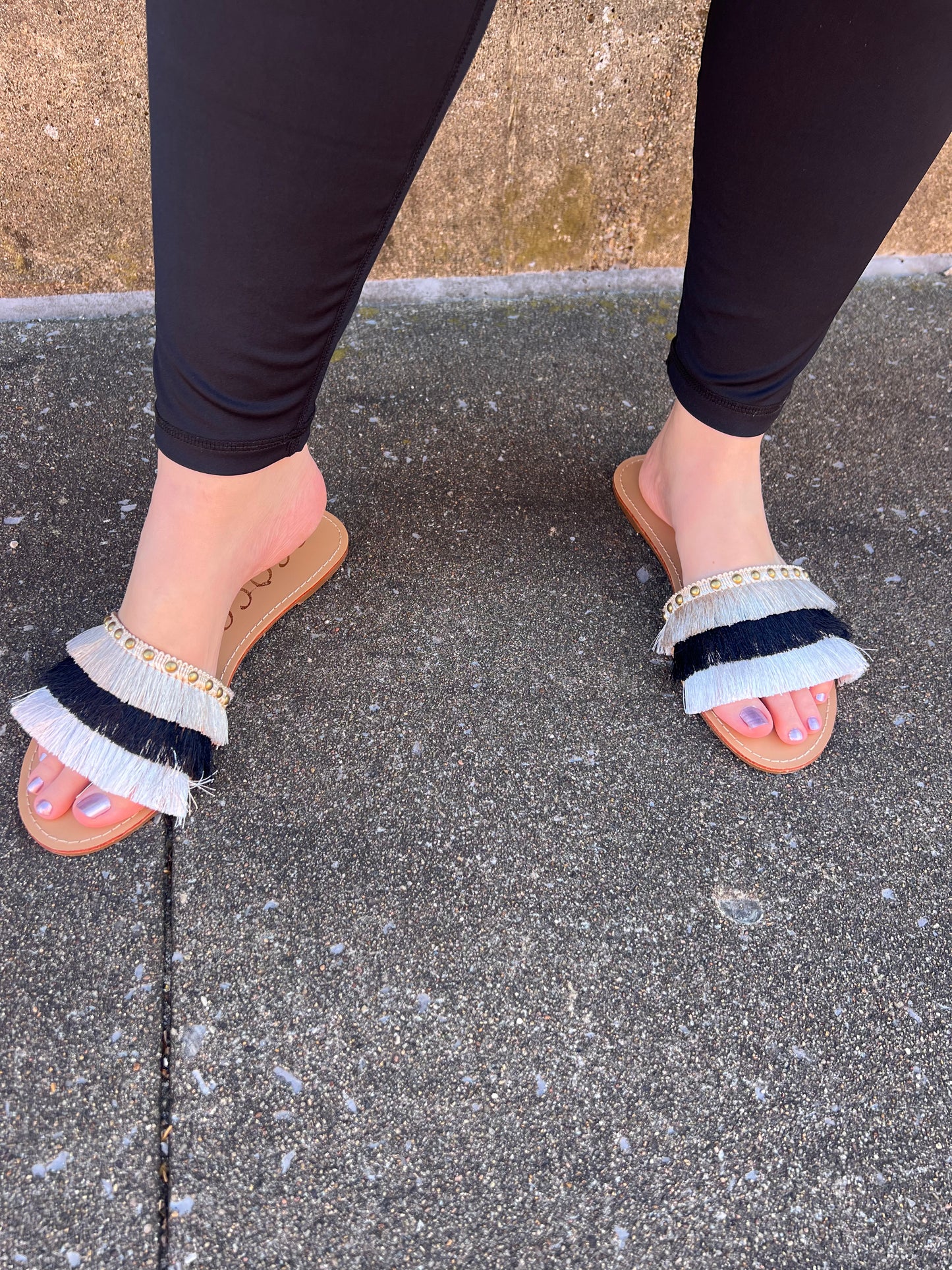 A pair of flat sandals with fringe detailing on the straps, worn by a person standing on a textured ground.