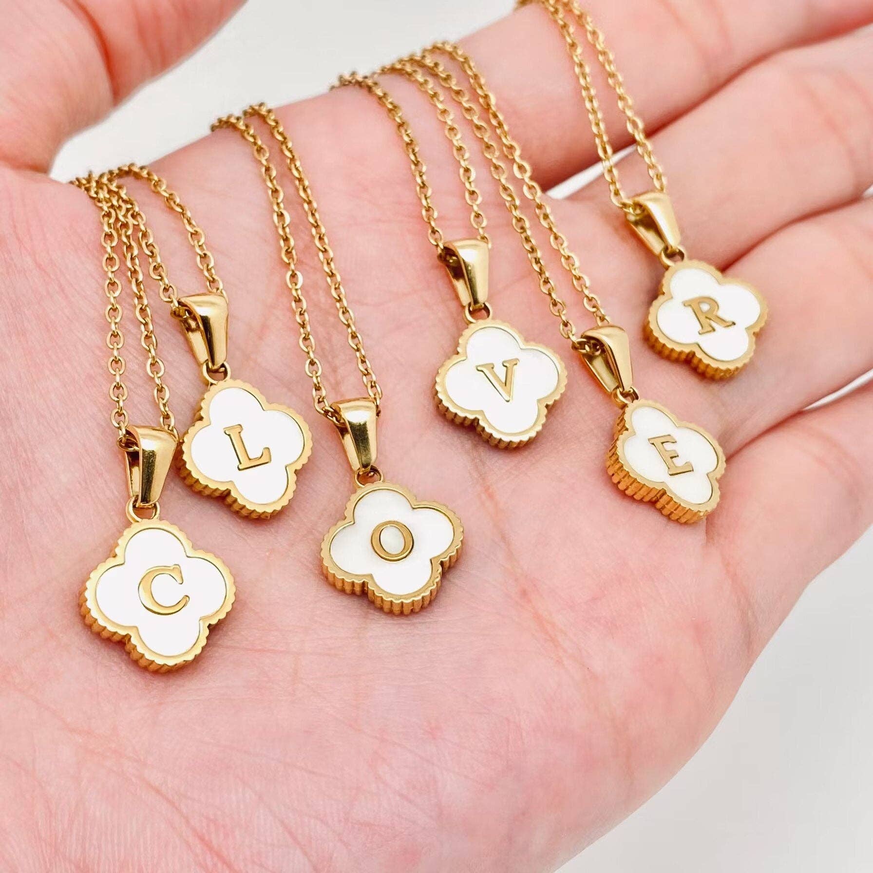 Gold necklaces with clover-shaped pendants displaying letters on a hand.