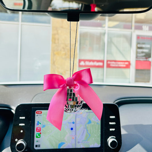 A pink bow disco car charm hanging from a vehicle's rearview mirror featuring a disco ball and pink ribbon.