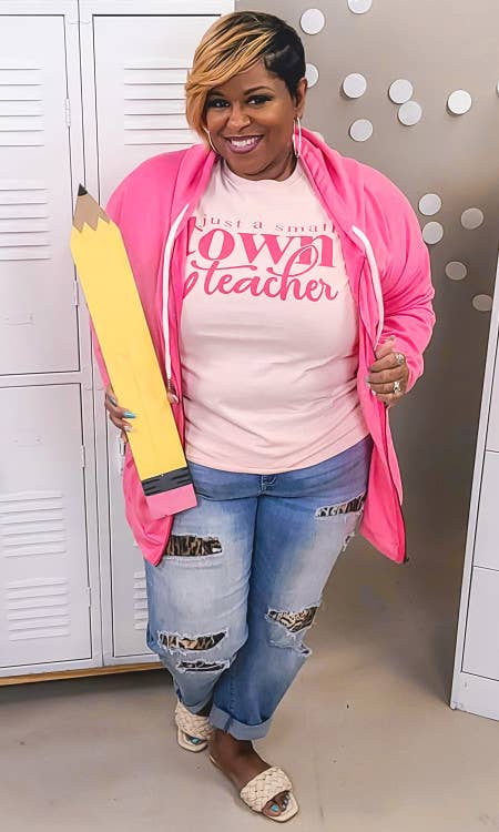 Person wearing a pink hoodie and 'just a small town teacher' shirt, holding a large yellow pencil.
