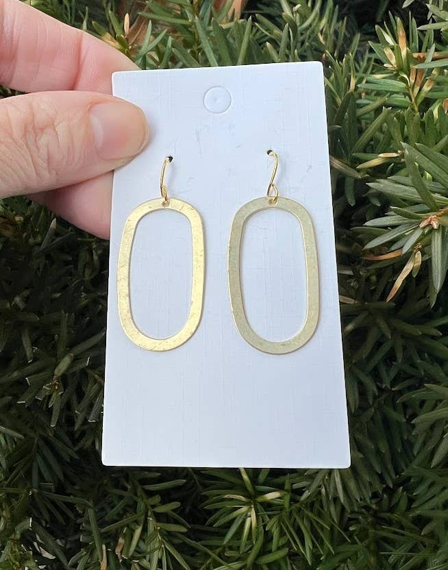 Gold oval earrings on a white card held by a hand with a green leafy background