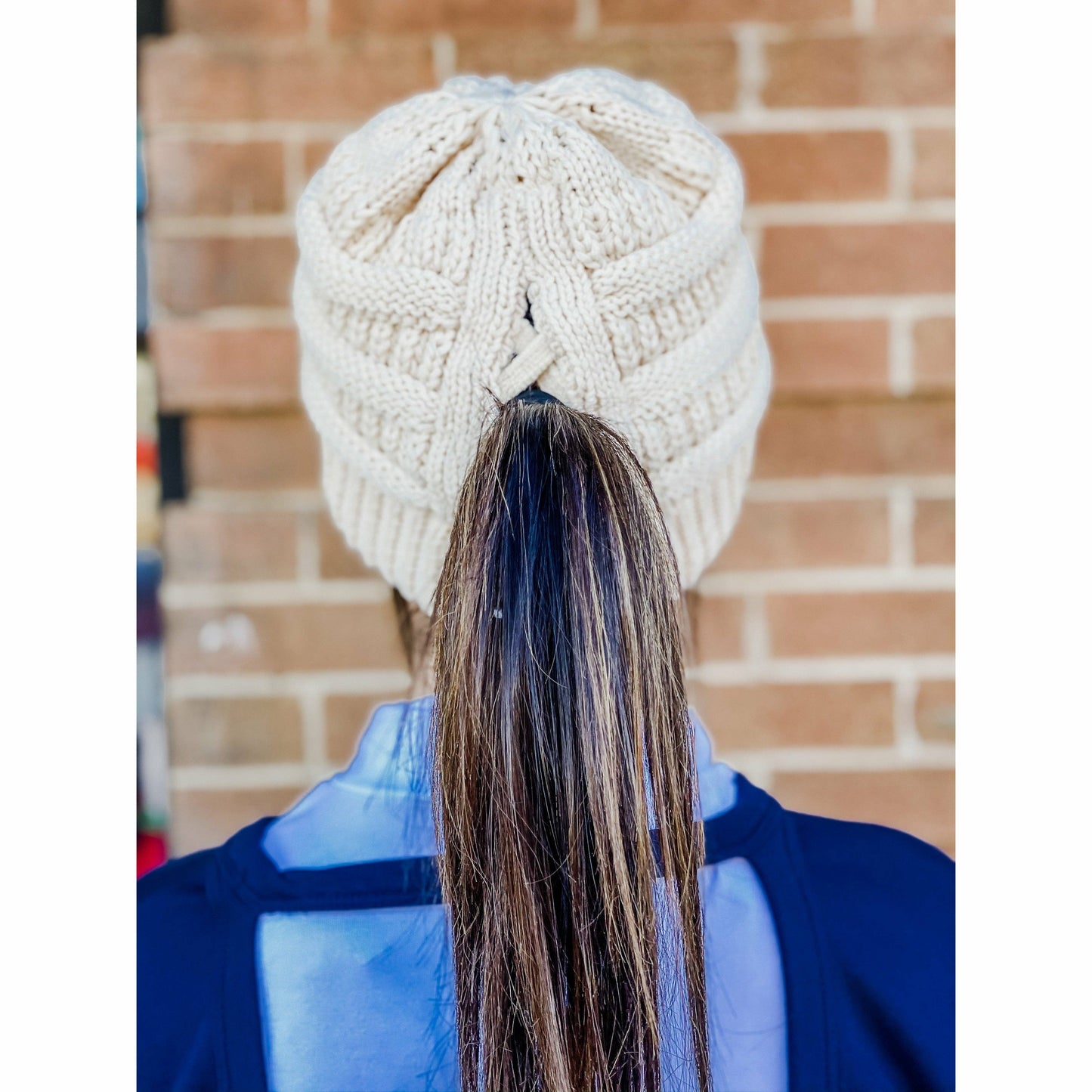 Person wearing a white knitted beanie with a textured pattern, standing against a brick wall.