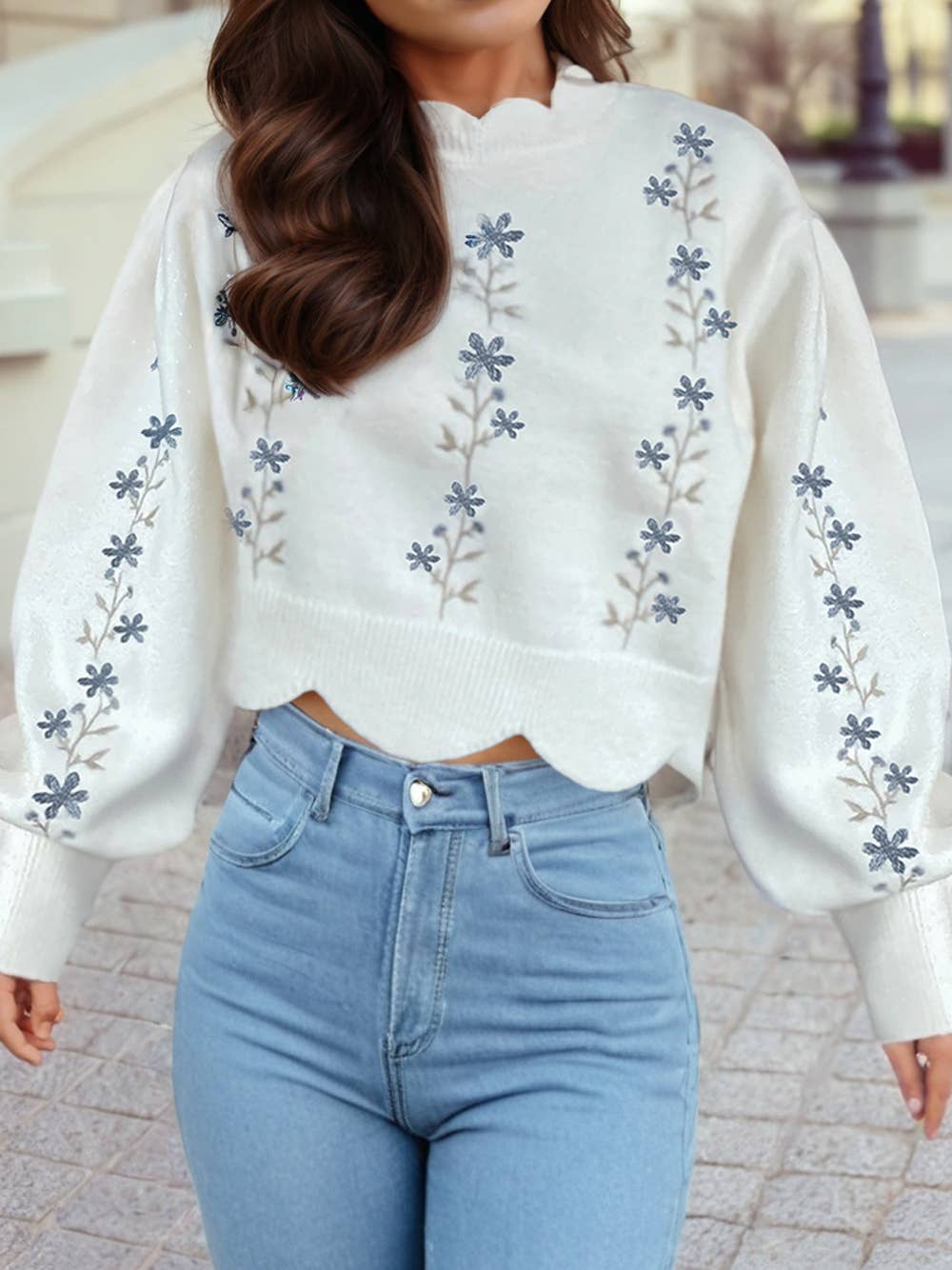 Person wearing a white sweater with floral embroidery and blue jeans outdoors.