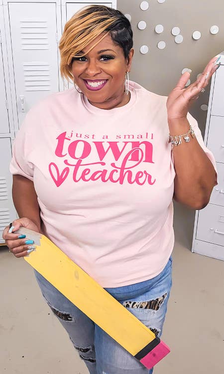 Woman wearing a pink 'just a small town teacher' shirt holding a yellow and pink sash.