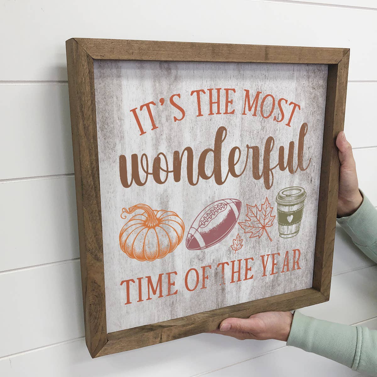 Decorative sign with autumn-themed text and illustrations held by a person against a white wall.