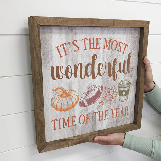 Decorative sign with autumn-themed text and illustrations held by a person against a white wall.