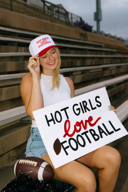A person wearing a red and white trucker hat with the text 'HOT GIRLS LOVE FOOTBALL' embroidered on it, sitting on a bench.