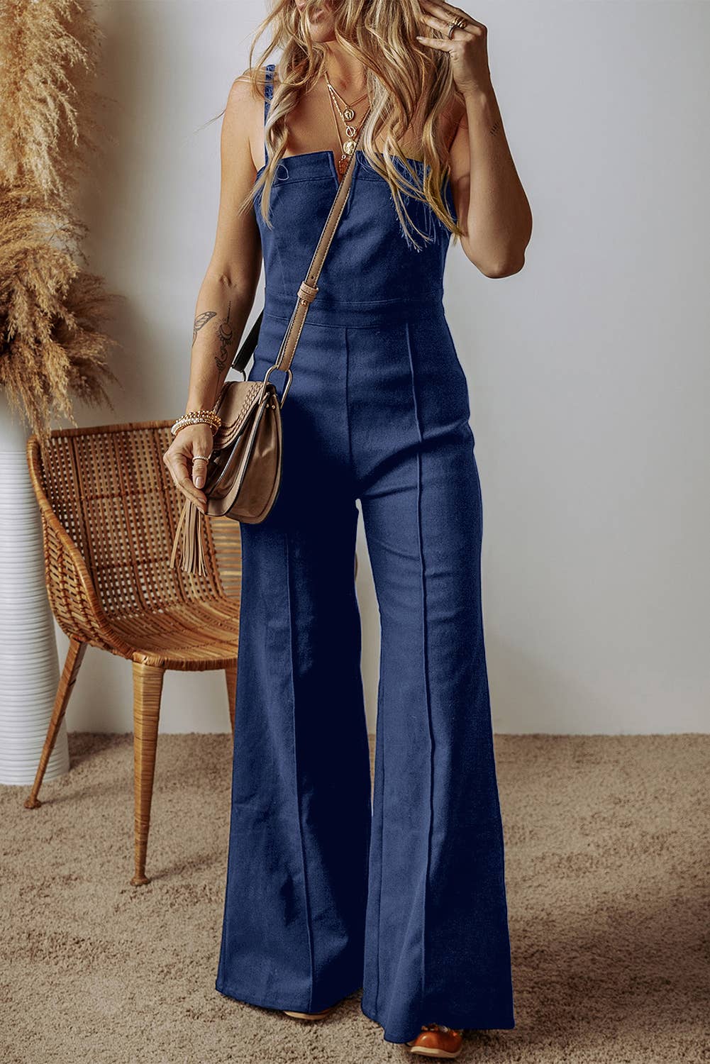A woman wearing a blue denim jumpsuit with spaghetti straps, a high waist, and flared legs. She has accessorized with a small handbag and is standing next to a wicker chair.