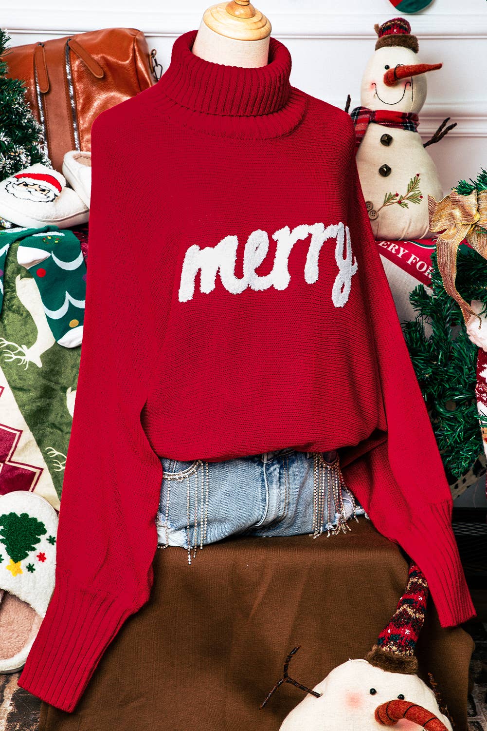 Red sweater with 'merry' text on a mannequin against a festive background with Christmas decorations.